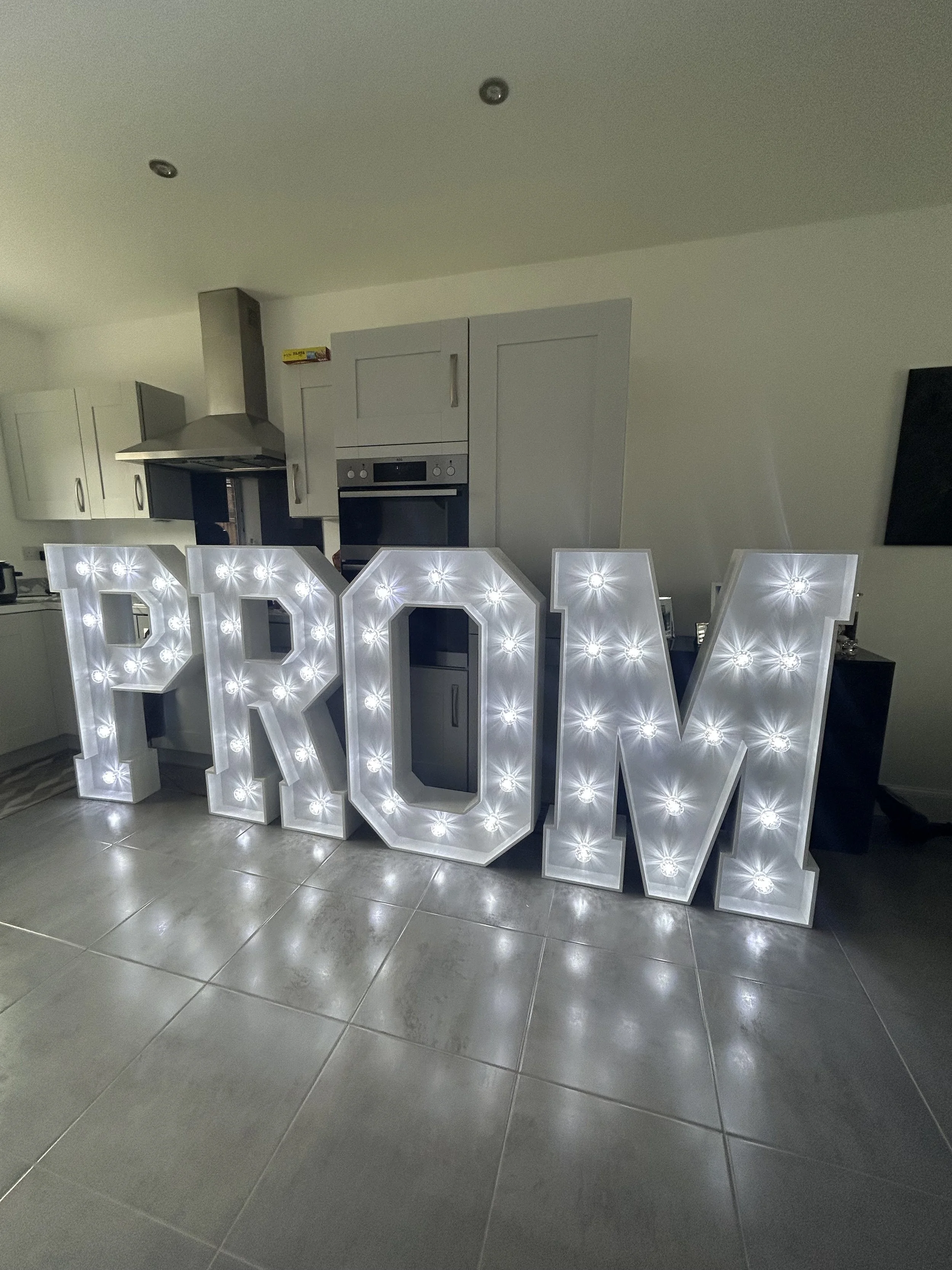 Illuminated marquee spelling out the word 'PROM' in a room with a kitchen background.