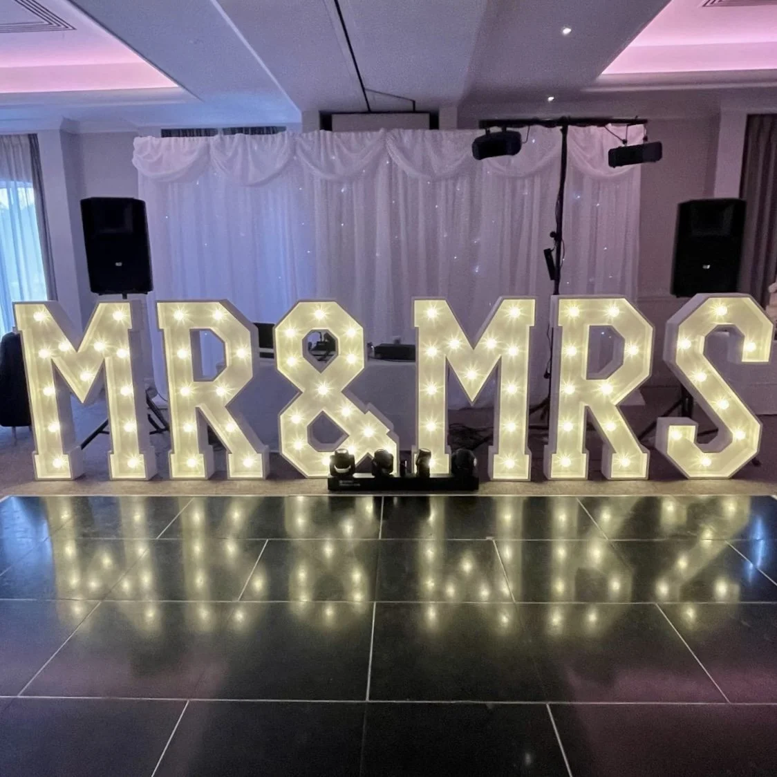 Illuminated marquee letters spelling 'MR & MRS' with a wedding reception setup in the background.