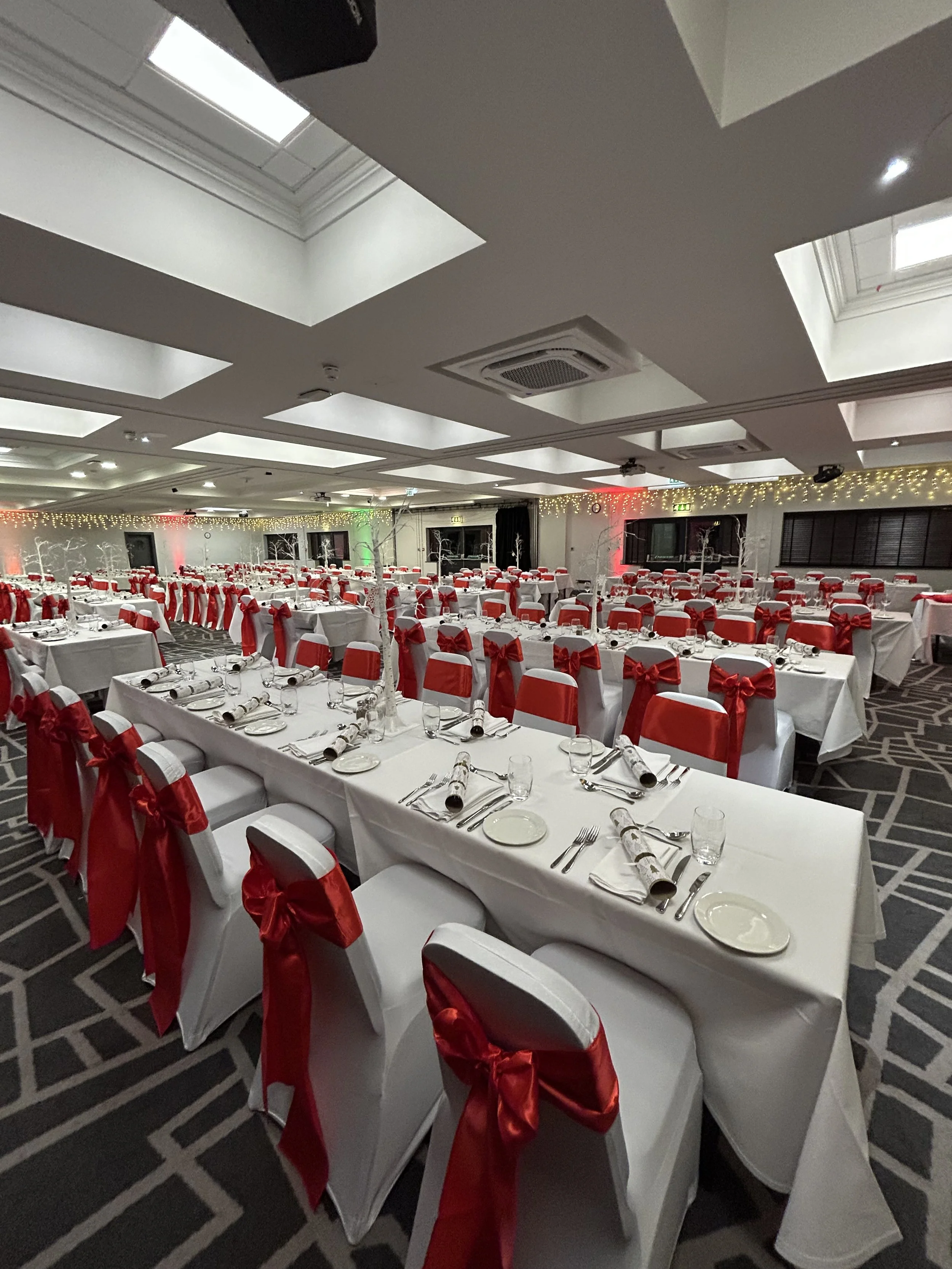 A banquet hall decorated for a Christmas celebration with long tables covered in white tablecloths, set with plates, glasses, and utensils. Chairs are adorned with white covers and red ribbons tied in bows. The room is decorated with string lights an