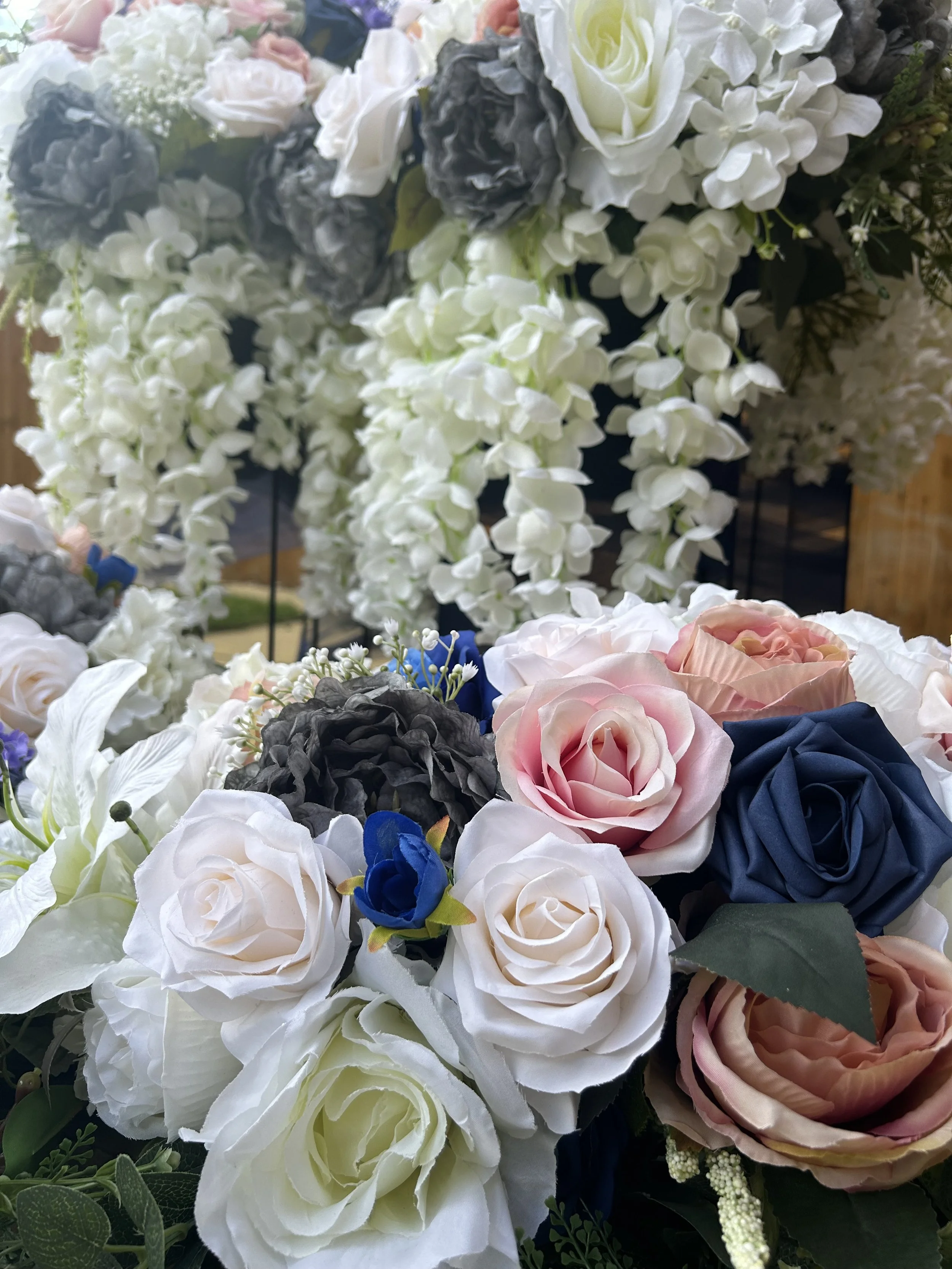 Close-up of a bouquet of artificial flowers including white roses, pink roses, blue flowers, and dark gray hydrangeas, with cascading white floral accents in the background.