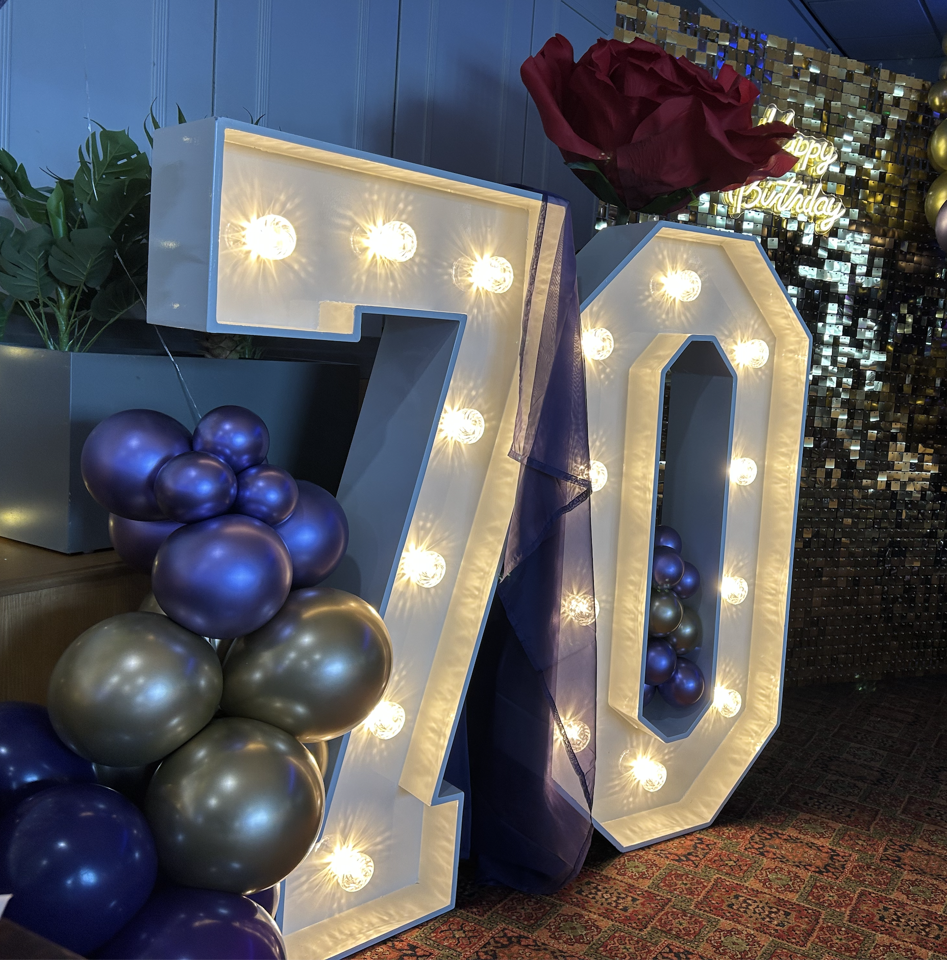 Large illuminated number 70 decorated with purple and gold balloons, a large red rose, and a 'Happy Birthday' sign in the background.