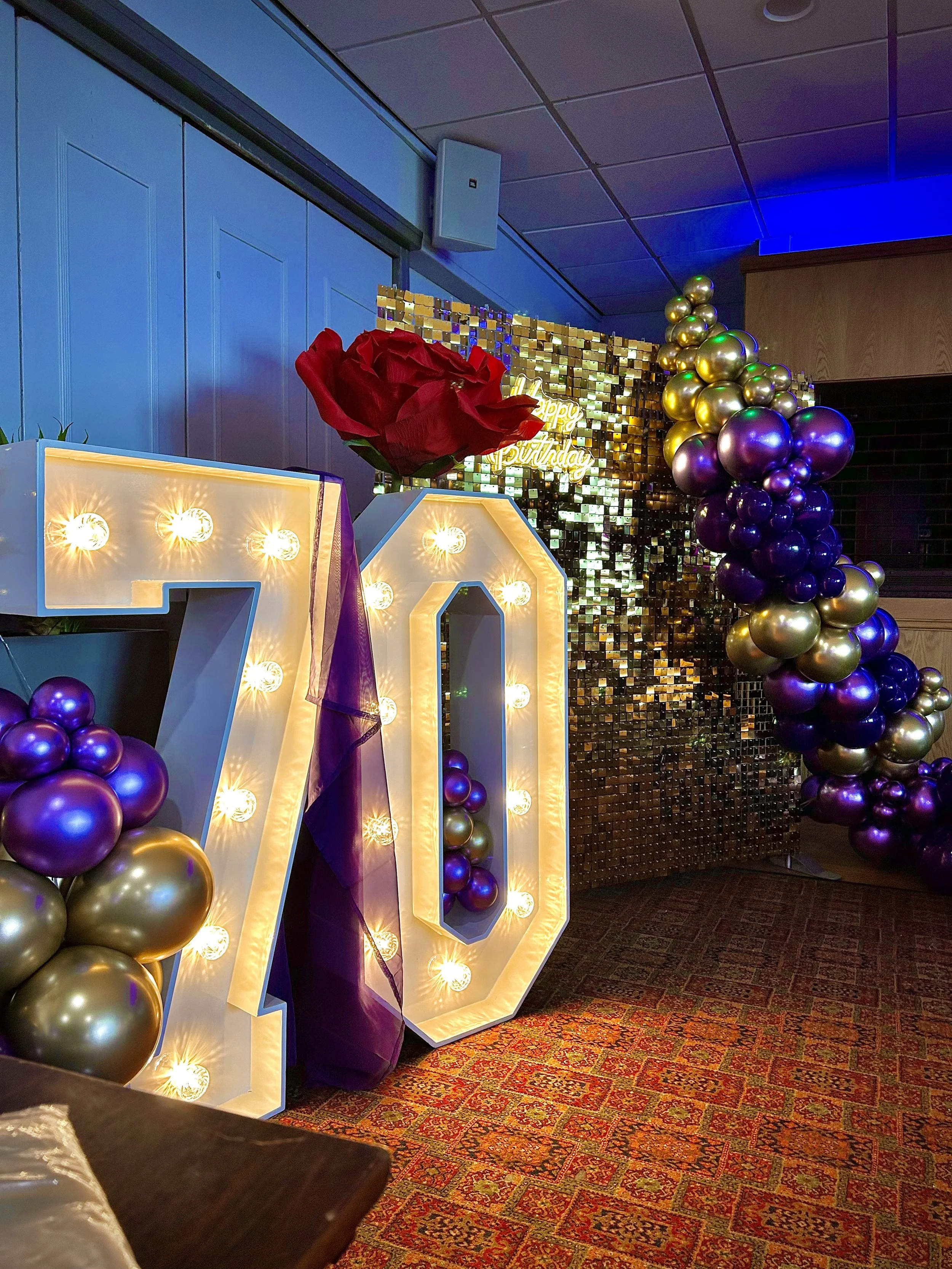 Decorative celebration setup with illuminated number '70', purple and gold balloons, a large red artificial rose, and a sparkling backdrop with a 'Happy Birthday' sign in gold.