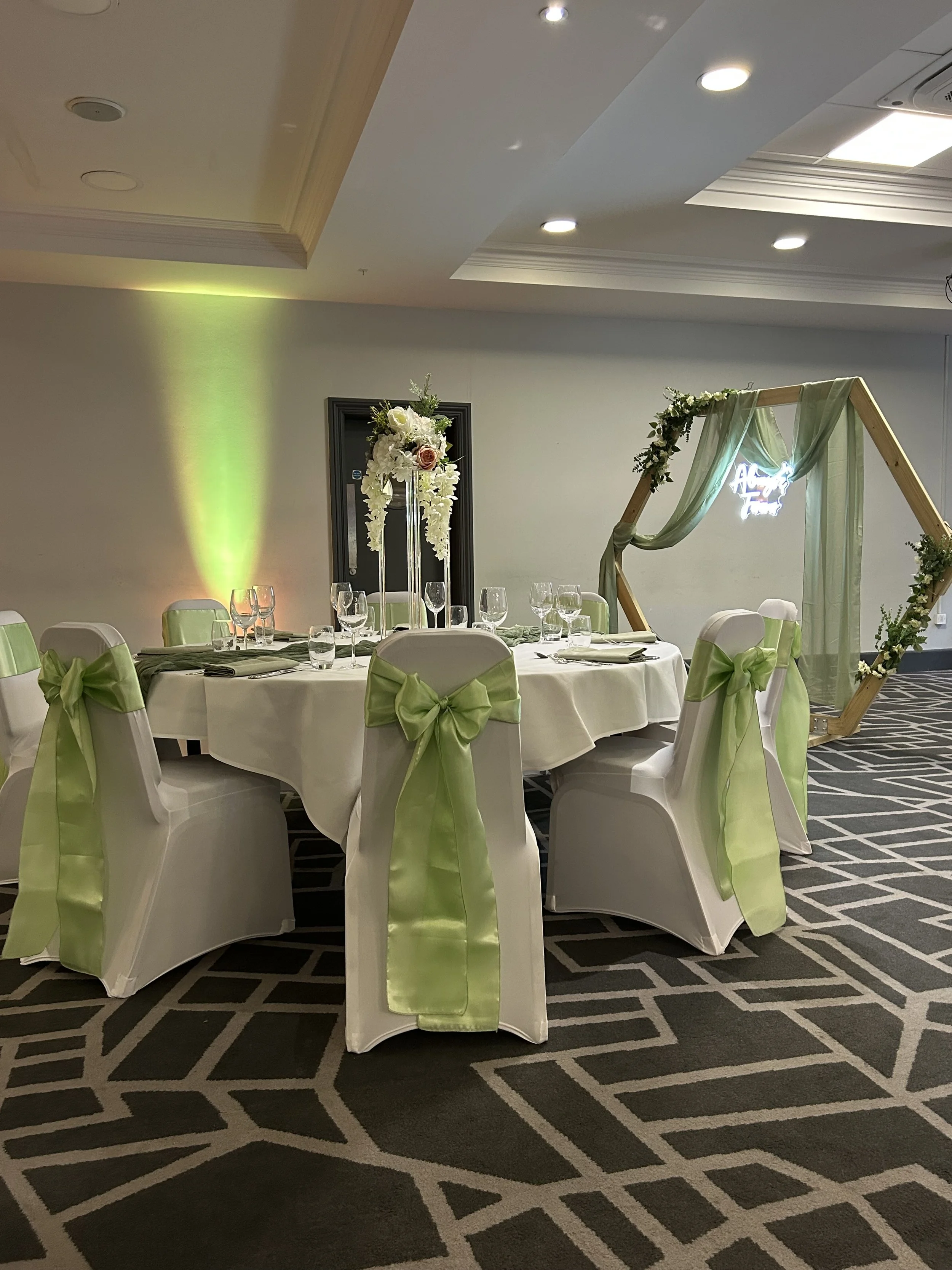 Graceful round table set for a celebration with white tablecloth, glassware, and white chairs decorated with lime-green ribbons, featuring a floral centerpiece and a geometric wooden arch with green drapes in a well-lit room.