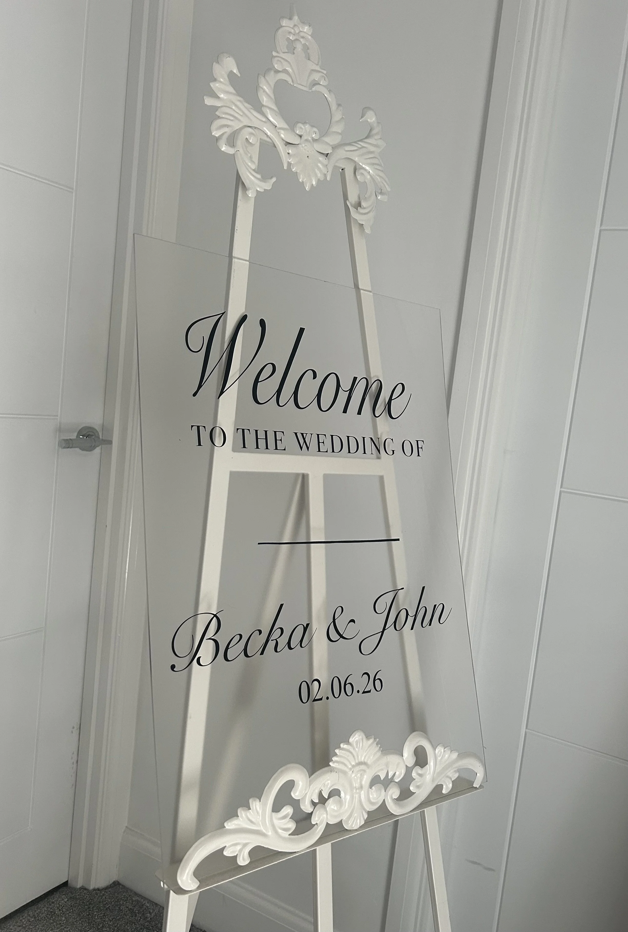 Wedding welcome sign on an easel with ornate white decorations, white walls, and a gray carpet.