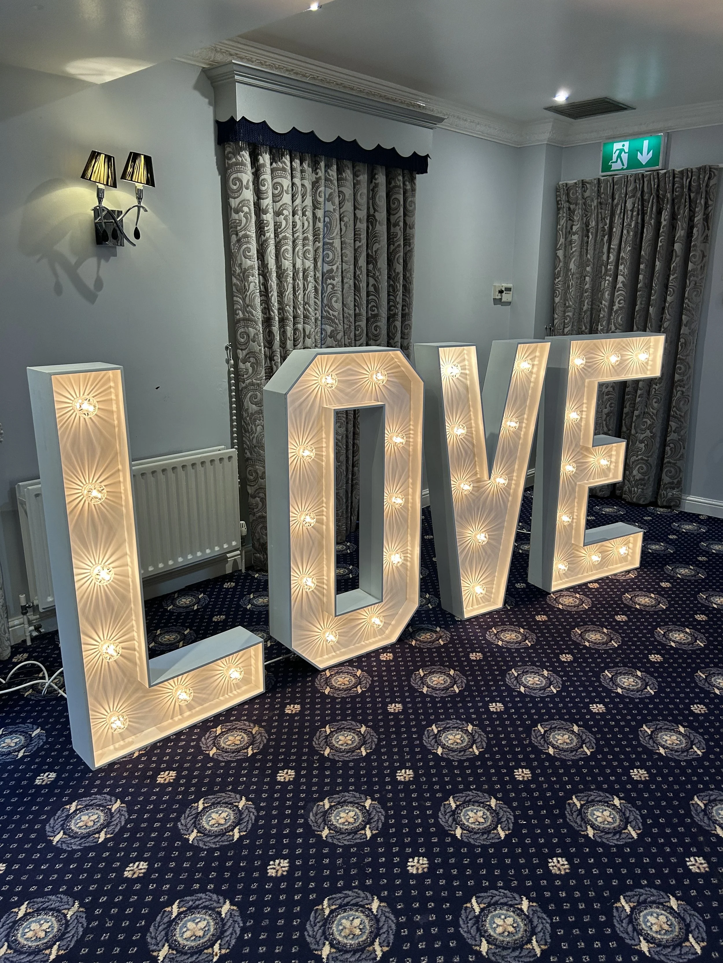 Large illuminated letters spelling 'LOVE' inside a room with patterned curtains, blue carpet, and wall-mounted lamps.