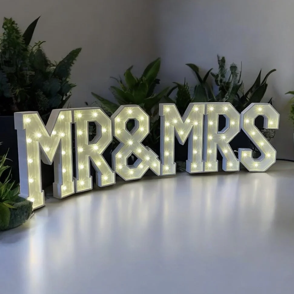 Decorative illuminated sign spelling 'MR & MRS' with white borders, placed on a white surface with green plants in the background.
