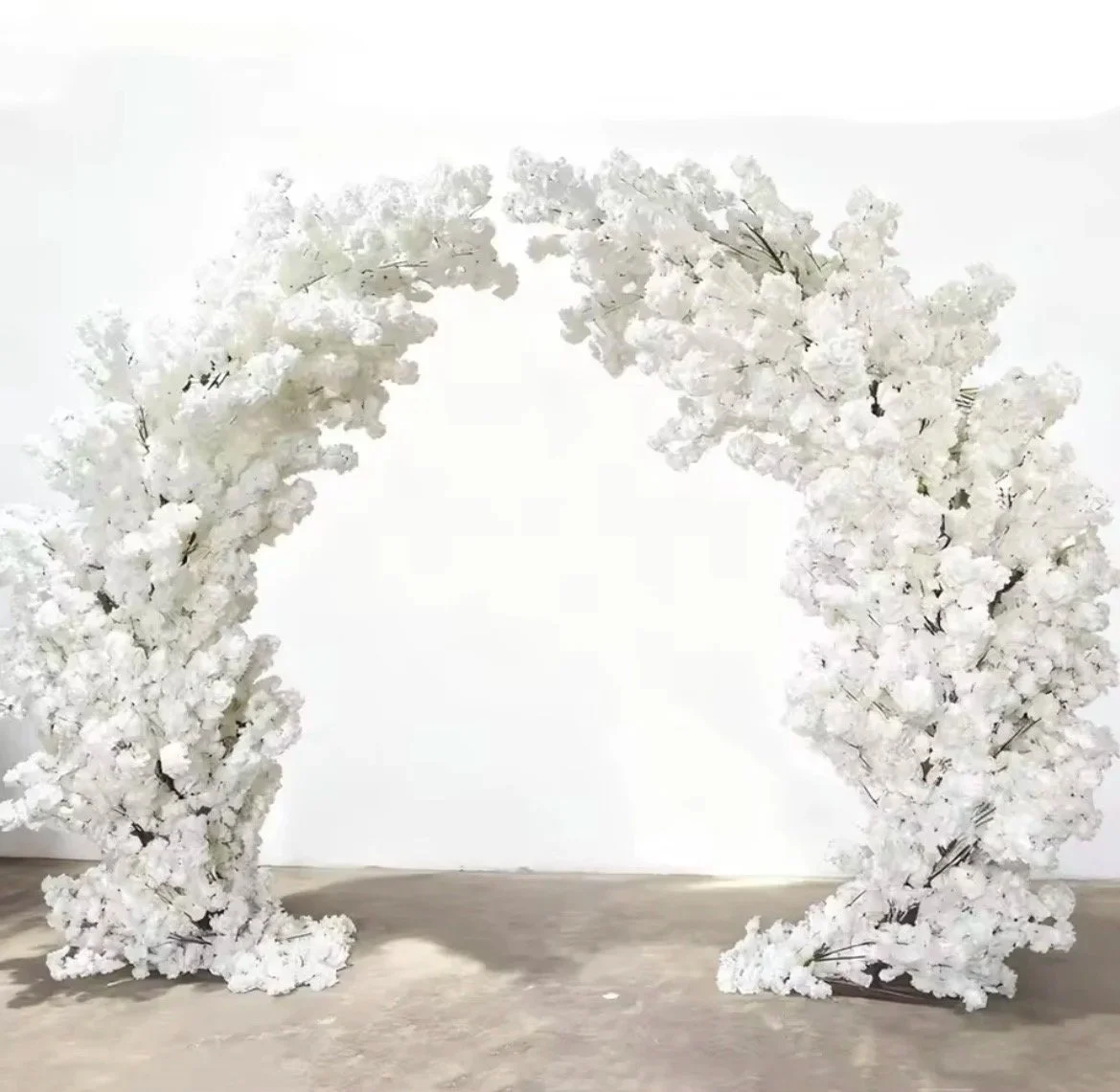 White floral arch made of artificial flowers and branches on a wooden floor against a white wall.