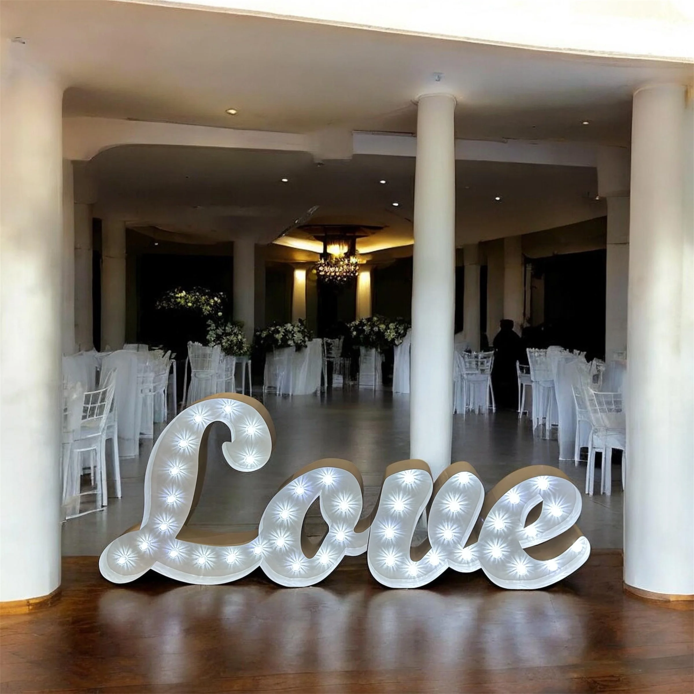 Large white decorative sign spelling 'LOVE' with illuminated bulbs, placed on a wooden floor, part of a decorated indoor event space.