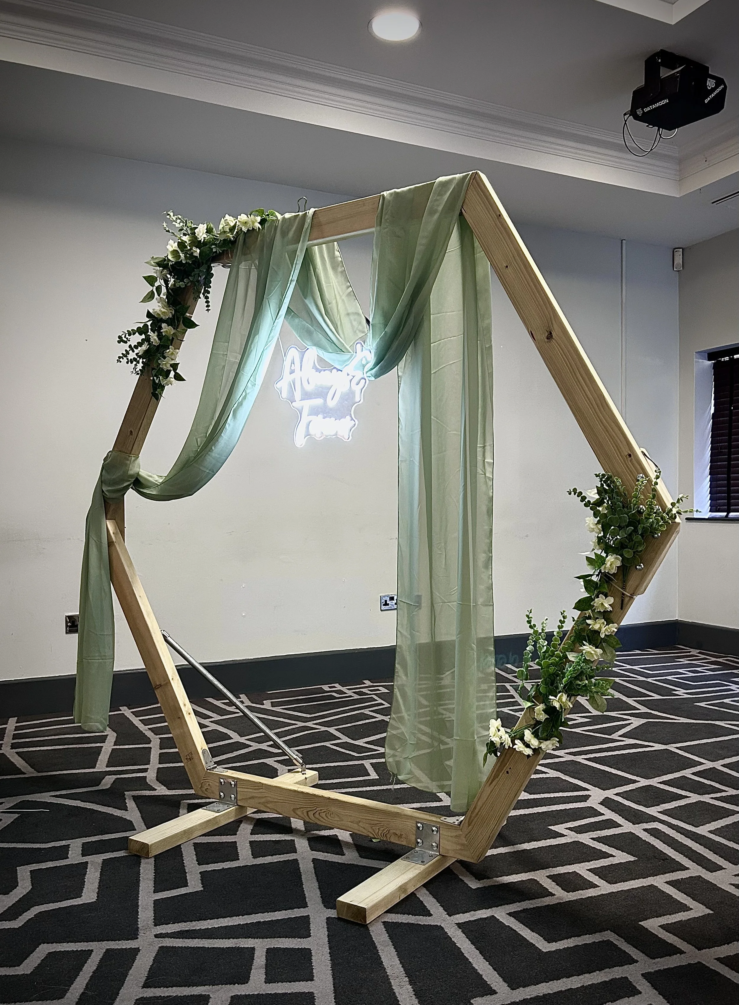 A wooden floral wedding arch decorated with greenery and light green sheer fabric, set up indoors on a patterned carpet.