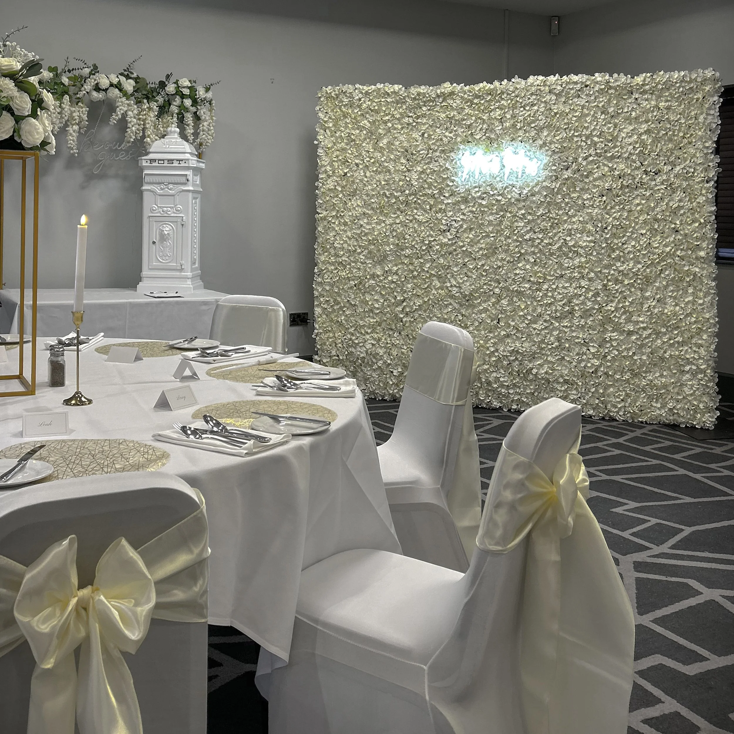A floral wedding backdrop with white flowers and a neon sign, decorated chairs with gold bows, a white post box, and dining tables set for a celebration.