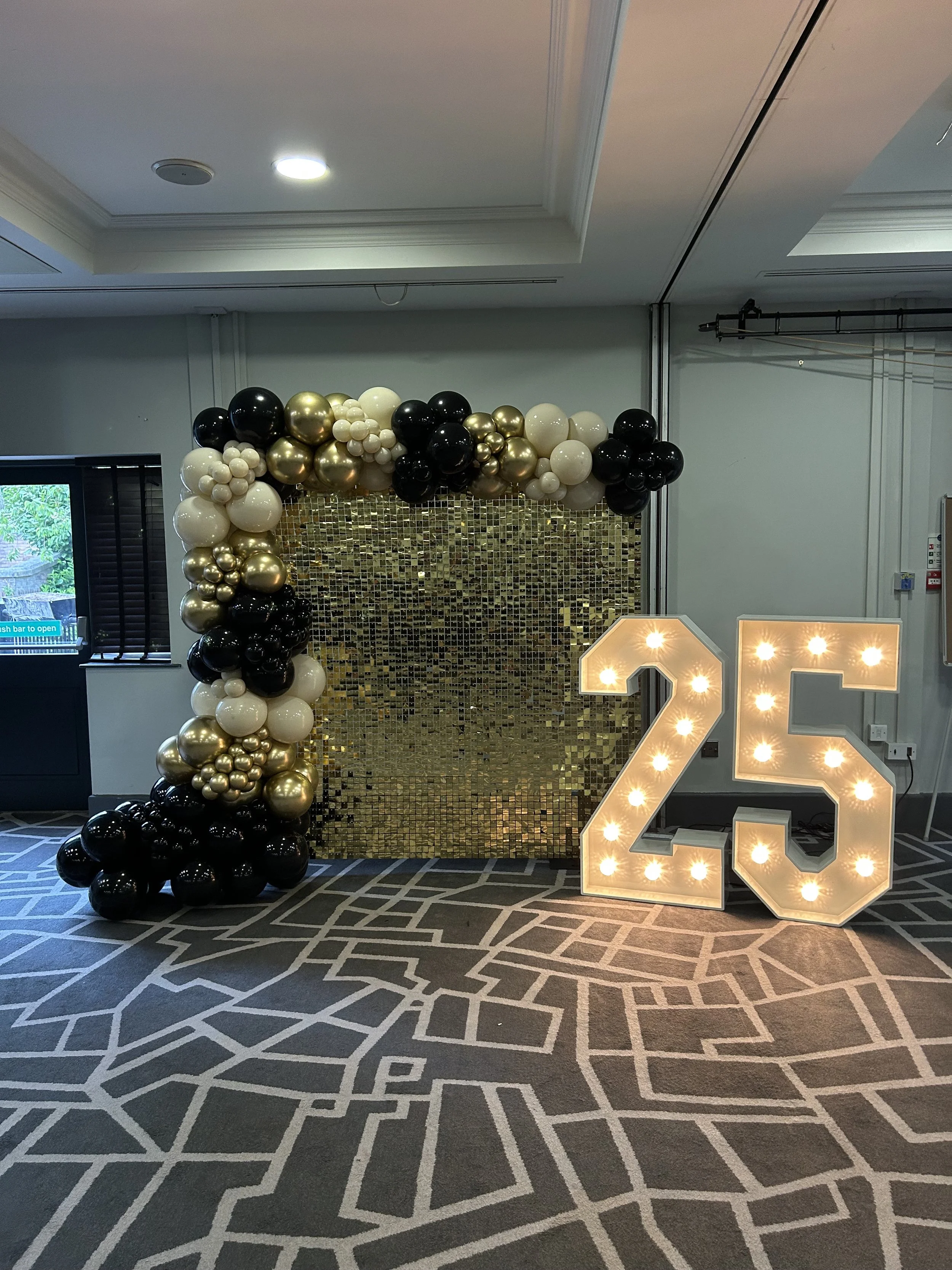 Decorative setup for a 25th birthday celebration featuring a balloon arch in black, white, and gold, a gold mosaic backdrop, and a large illuminated number 25.
