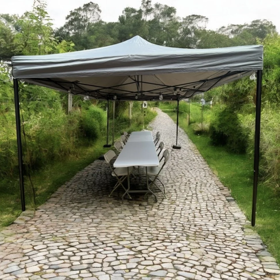 An outdoor dining setup with a white rectangular table surrounded by folding chairs under a gray canopy tent on a cobblestone pathway, with greenery and trees in the background.