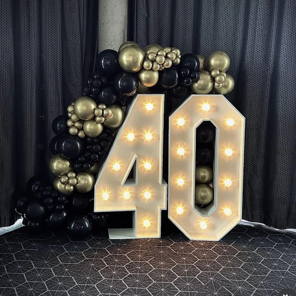 Decorative birthday display with large illuminated number '40' and black and gold balloons arranged around it against a dark curtain background.
