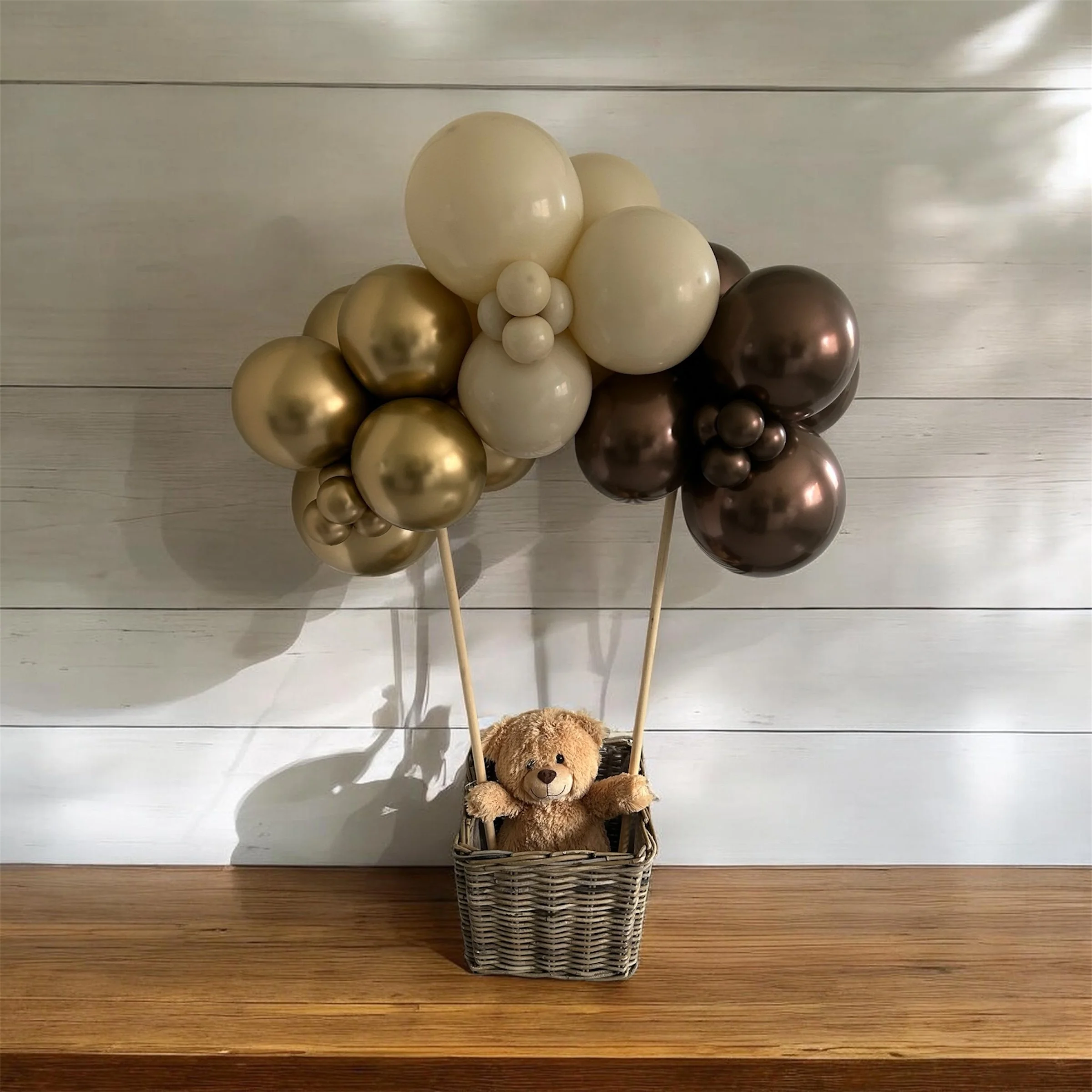 A collection of balloons in shades of gold, cream, and brown, tied to sticks and placed in a woven basket with a teddy bear, against a white wooden wall background.