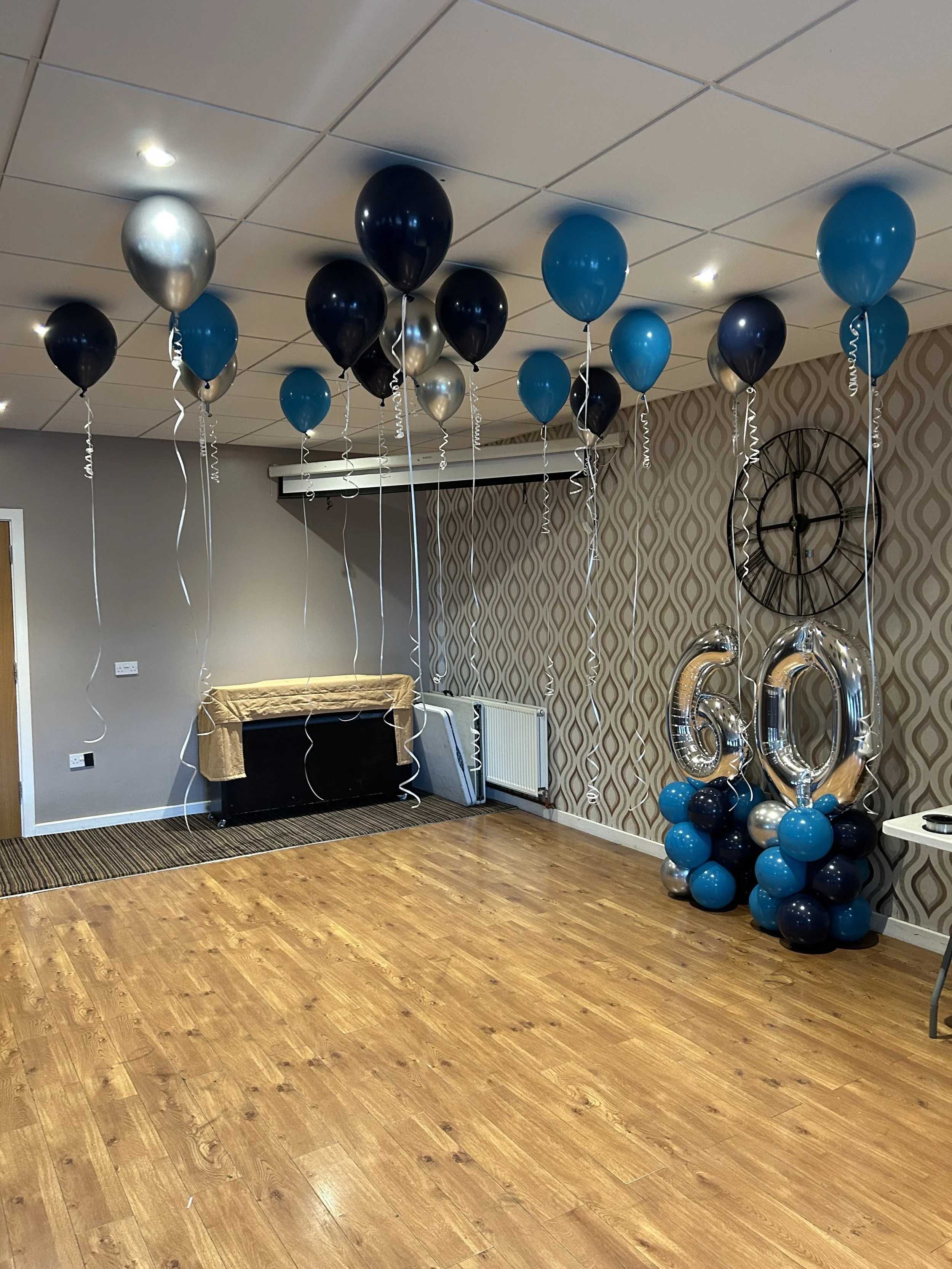 Room decorated for a 60th birthday celebration with black, blue, and silver balloons, including a cluster of balloons shaped as the number 60, and a wooden floor.