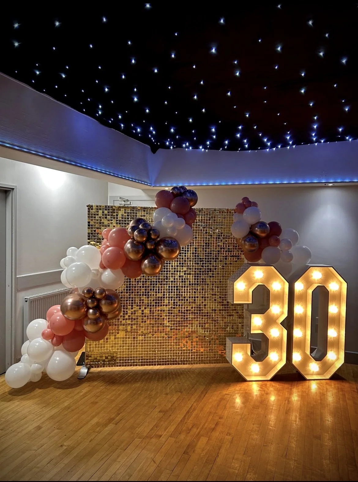 Decorations for a 30th birthday celebration with a large light-up number '30', a gold mosaic wall backdrop, and an arch of balloons in white, pink, and metallic rose gold colors. The ceiling has star-like fairy lights, and the venue features wooden f