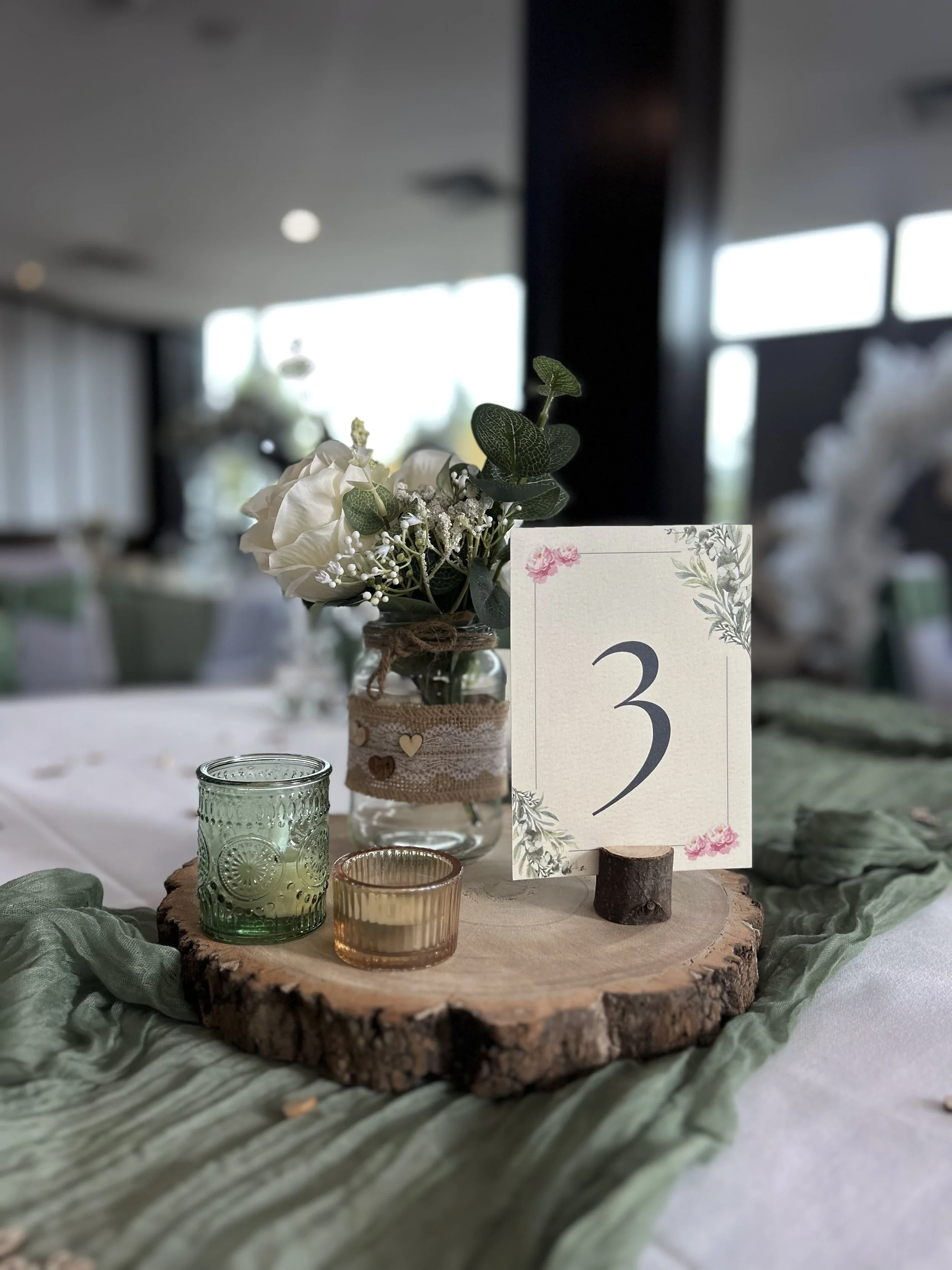 Centerpiece with white flowers and green leaves in a small jar wrapped with burlap and heart-shaped embellishments. Surrounding it are small, colored glass candle holders in green, amber, and pink on a round wooden slice. A table number card with floral design and the number 3 is placed on a small wood stand.
