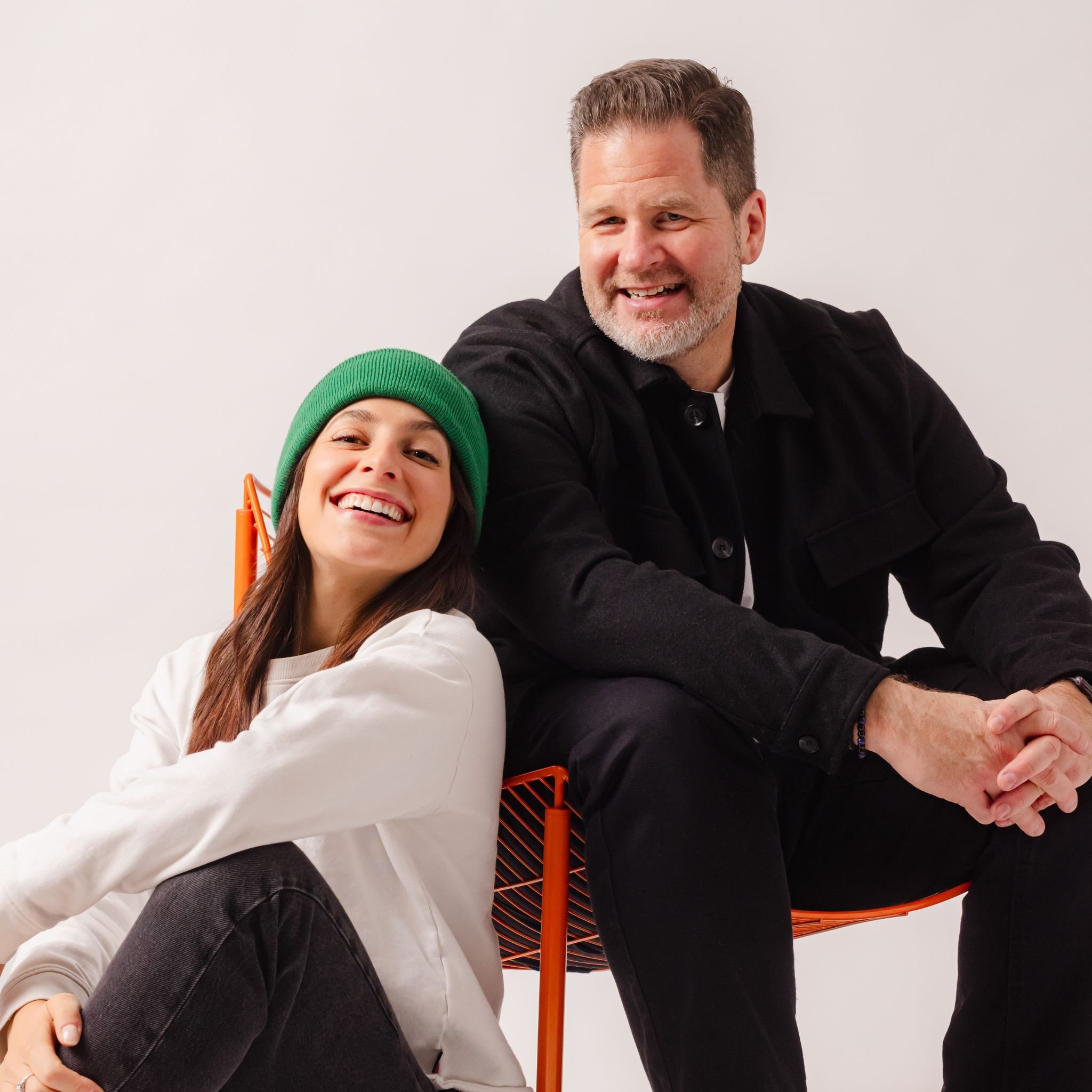 A man with gray hair and beard smiling, sitting next to a young girl with a green hat and white sweatshirt, both smiling, with a white wall background.