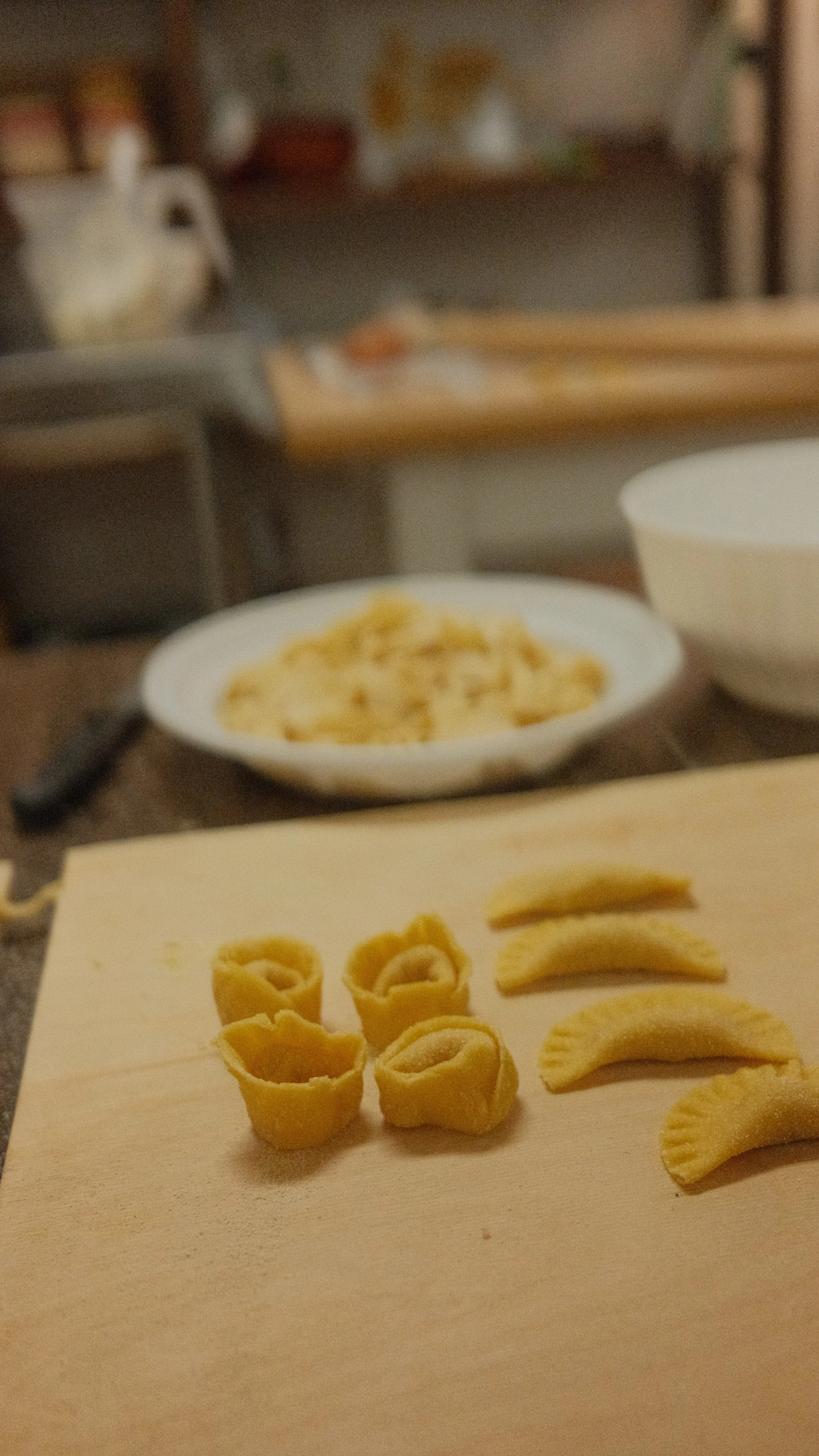 Uncooked pasta shapes including small rolled versions, half-moon, and straight pasta pieces on a wooden cutting board in a kitchen setting.