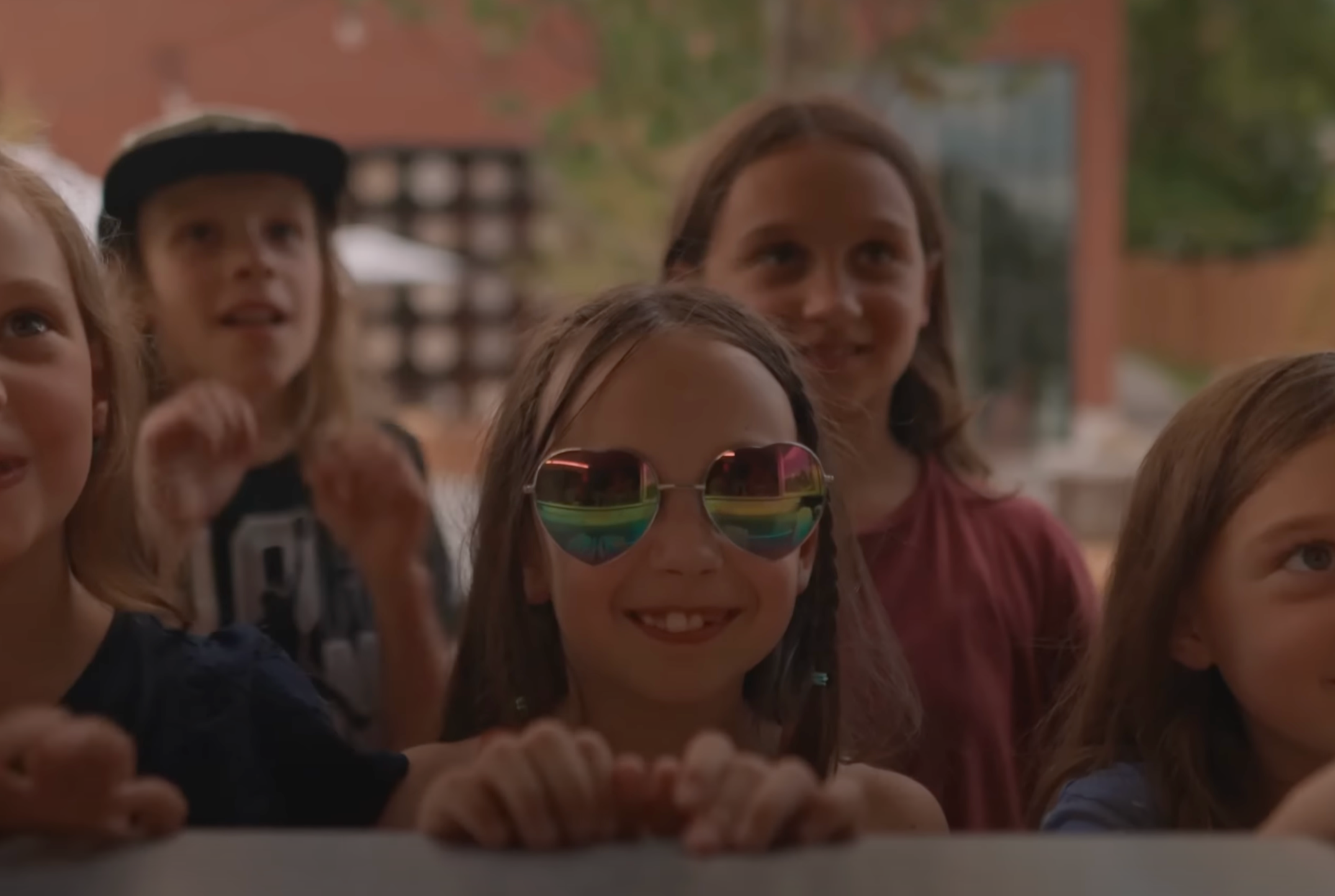 Group of children and a woman smiling and looking up, with some wearing sunglasses and one child wearing a black cap, outdoors with trees and a building in the background.