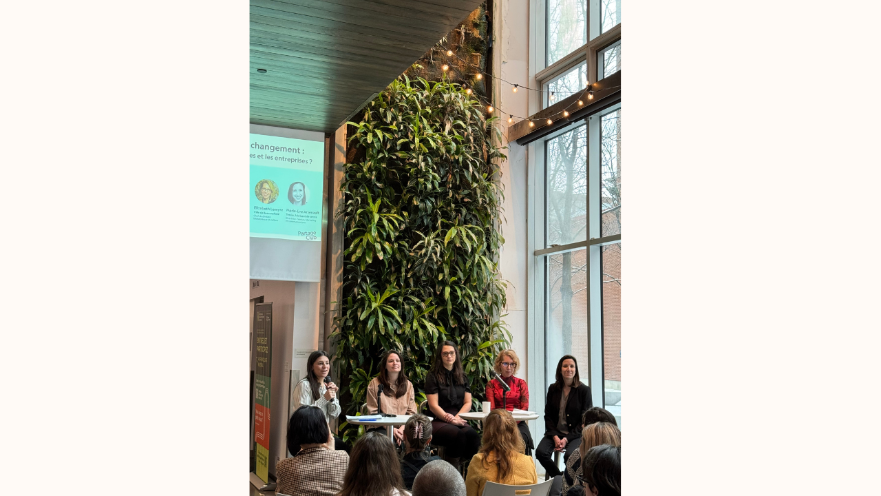 A panel of five women sitting at a table in front of an audience. A large green plant wall is behind them, and a presentation slide is projected on the wall to the left. The room has tall windows letting in natural light and string lights hanging overhead.