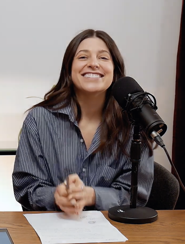 A woman with long brown hair, smiling and clapping in front of a microphone during a recording or podcast session.