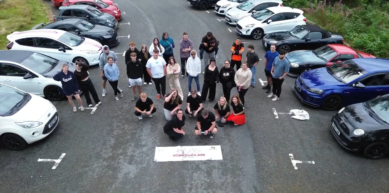 Group of people standing and sitting in a parking lot, surrounded by cars, with a banner on the ground in front of them.