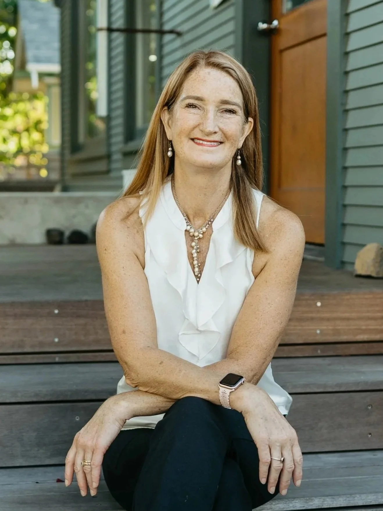 A woman with long hair, wearing a white sleeveless blouse, black pants, and black sandals, sitting on wooden steps outside a house, smiling at the camera.