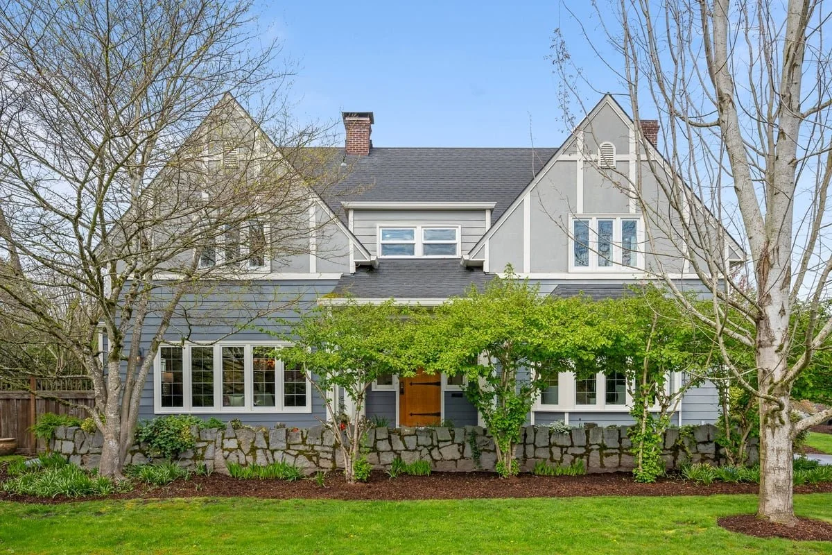 A large gray house with a stone foundation, multiple windows, a steep roof, and a central wooden front door. There are trees and well-maintained grass in front of the house.