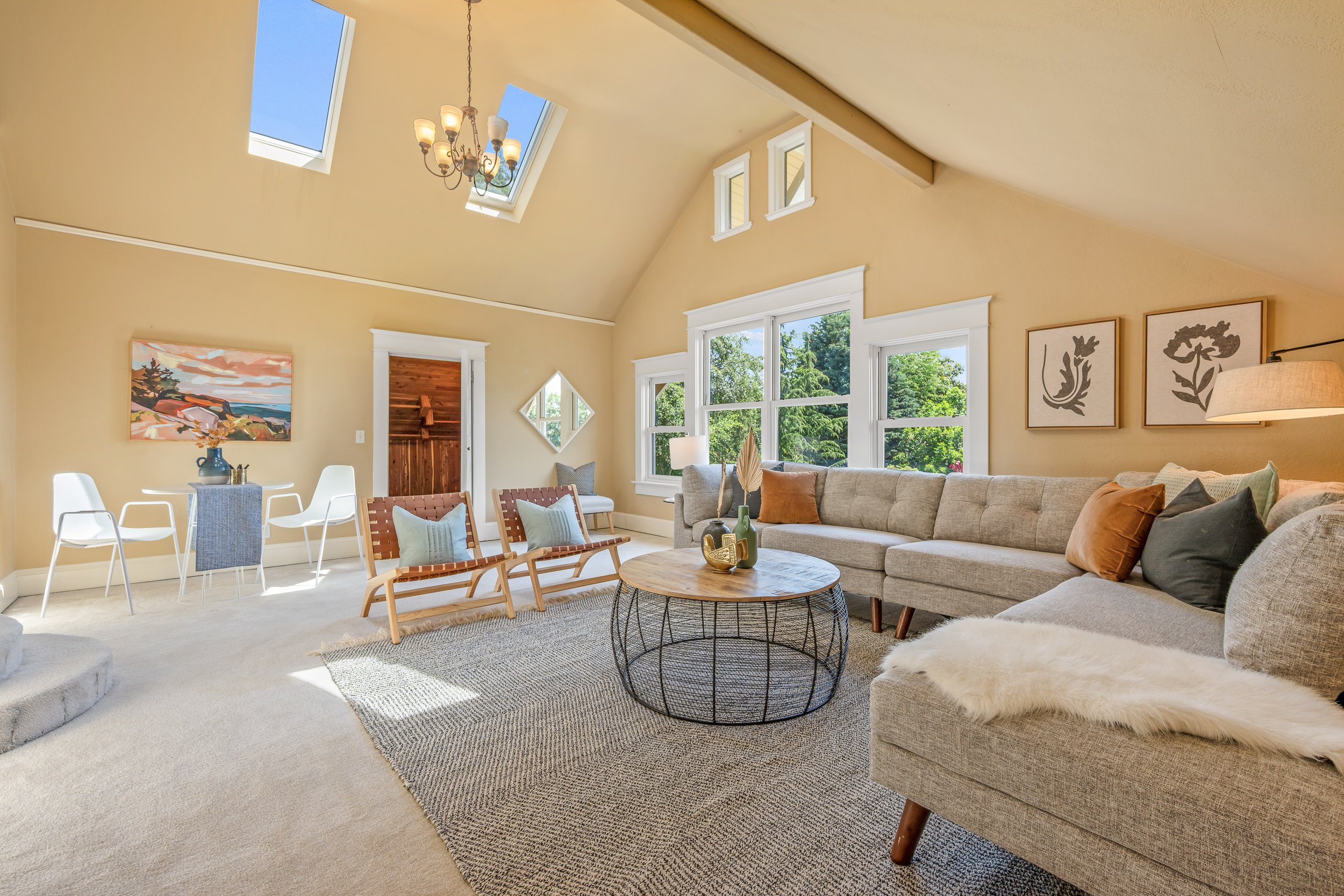 Spacious living room with beige walls, large windows, and skylights. Features a light grey sectional sofa with throw pillows, a round coffee table, and a rug. Decorative wall art, a chandelier, and a side table with a lamp. Bright and airy with greenery visible through the windows.