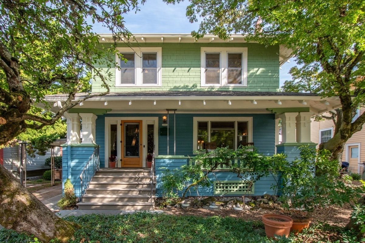 Colorful two-story house with light green upper section and blue lower section, front porch with steps, trees, greenery, and potted plants.