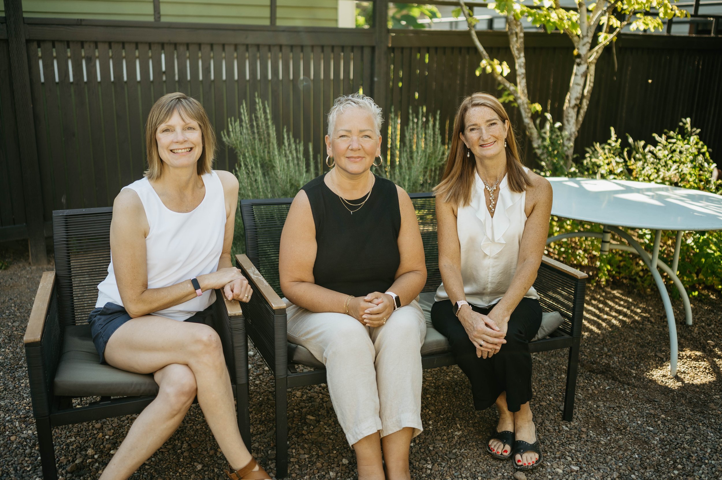 Three women sitting on a black outdoor bench in a garden, smiling at the camera.