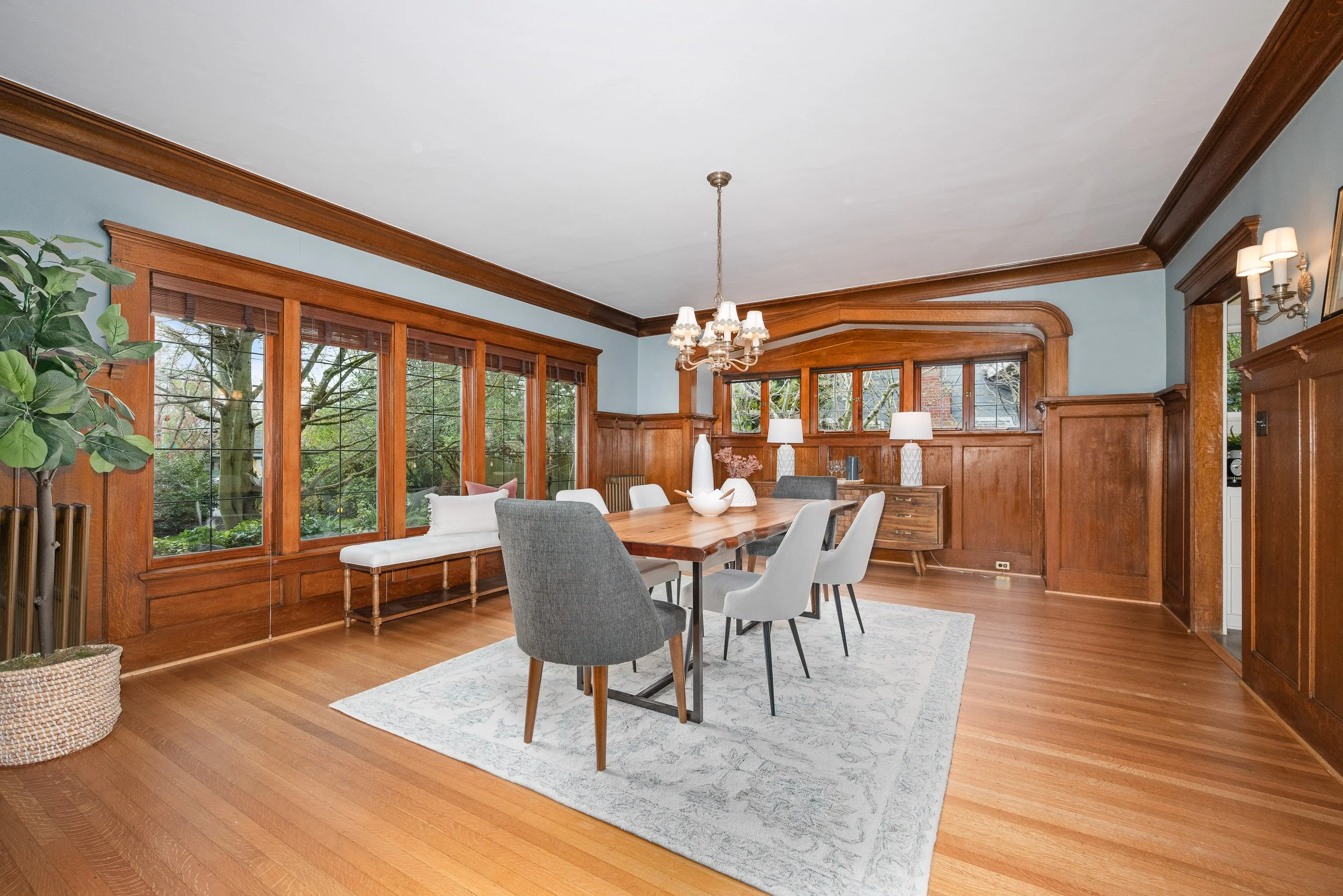 A dining room with large windows, wood paneling, and hardwood floors. The room features a wooden dining table surrounded by mixed gray and white chairs, a bench by the window, and a rug underneath the table. There are lamps and decorative items on side tables and a chandelier hanging from the ceiling.