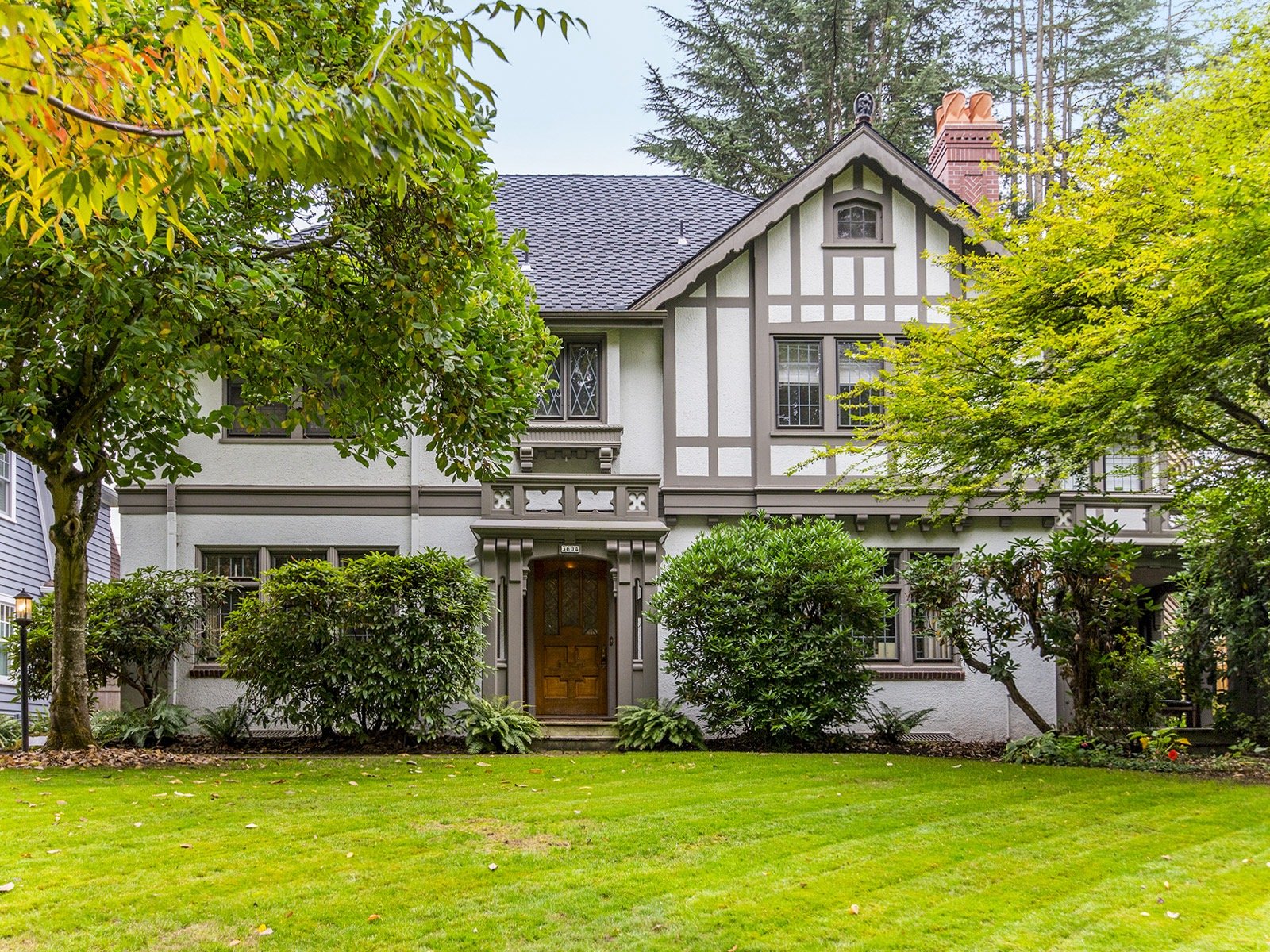 A large, two-story house with a Tudor-style design, featuring a wooden front door, decorative half-timbering, and multiple windows. The house is surrounded by a well-manicured lawn and lush green trees and bushes.