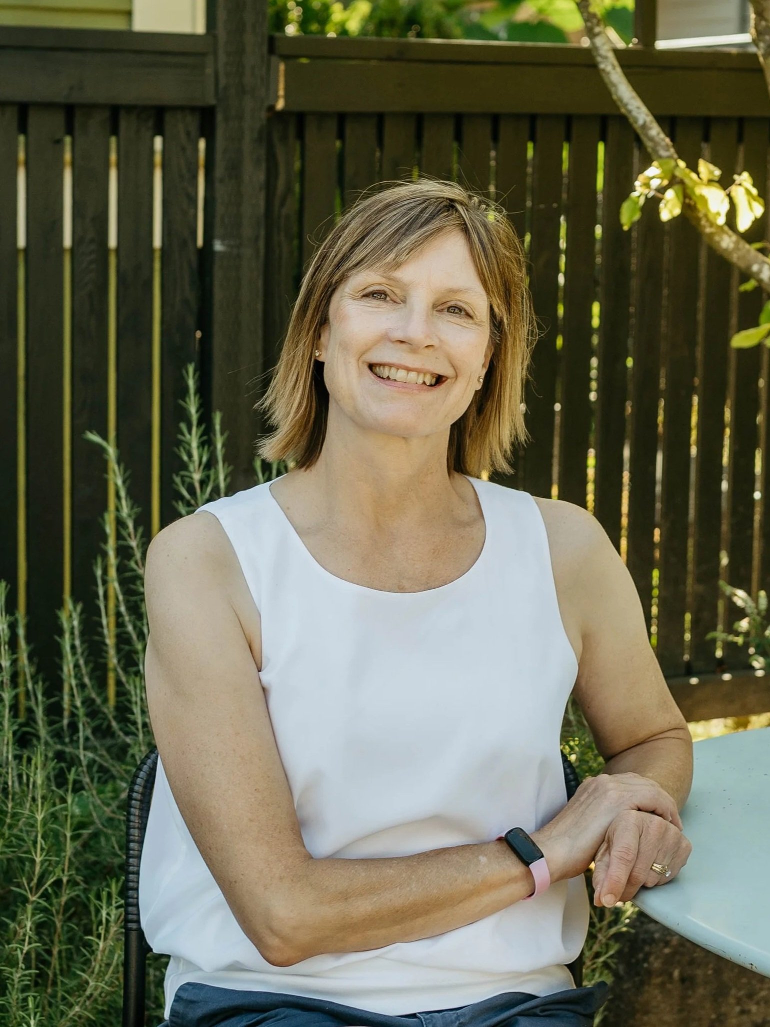 A woman sitting outdoors at a round table, smiling, with a garden and wooden fence in the background.