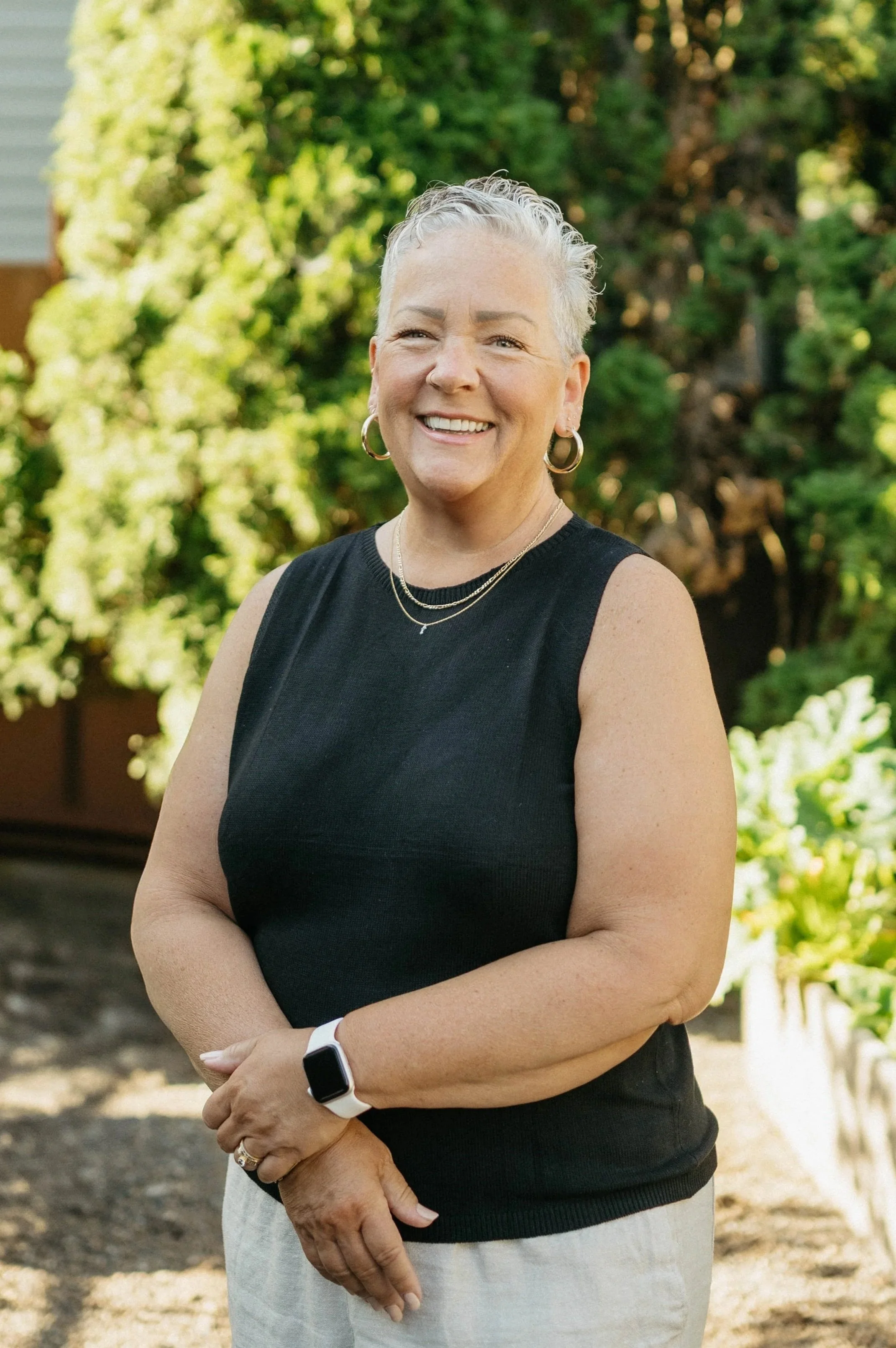 A smiling older woman with short gray hair, wearing a black sleeveless top, earrings, necklaces, and a smartwatch, standing outdoors in front of green shrubbery.