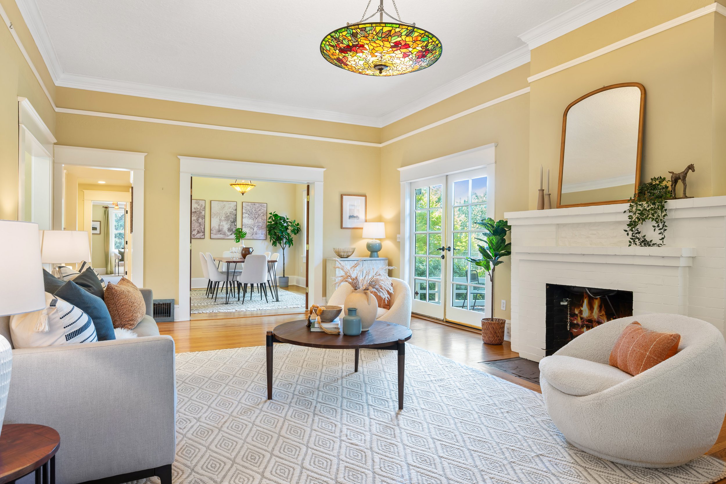 Bright living room with cream walls, white fireplace, glass doors, and a colorful stained glass ceiling light fixture. Decor includes potted plants, vases, and a patterned rug.