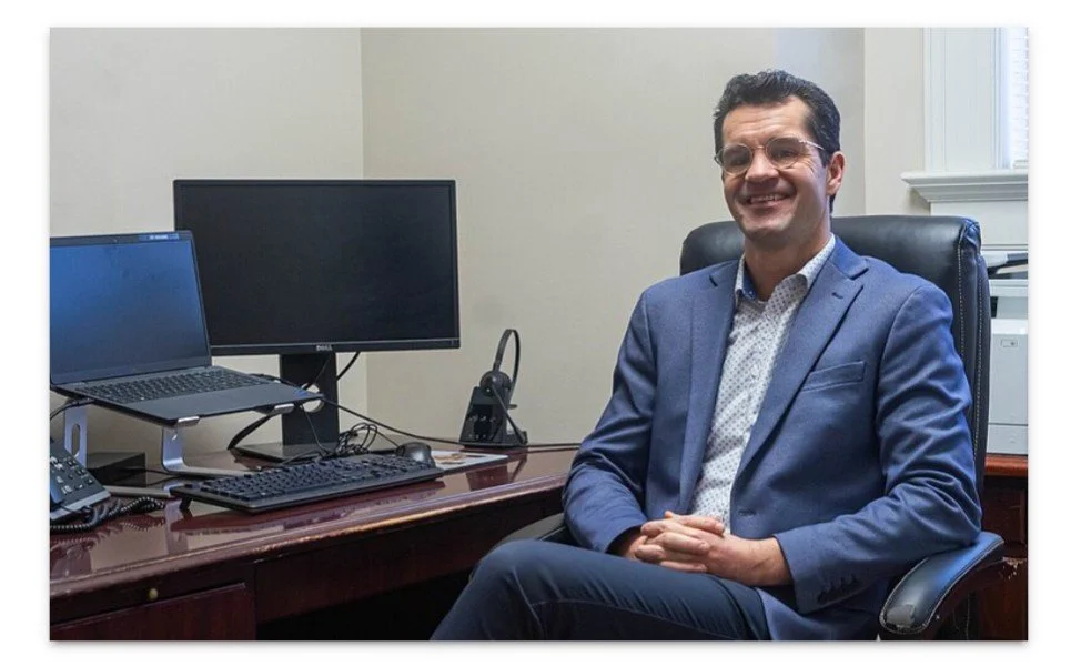 A man sitting at a desk in an office, smiling, with two computer monitors, a keyboard, a telephone, and a landline headset on the desk.