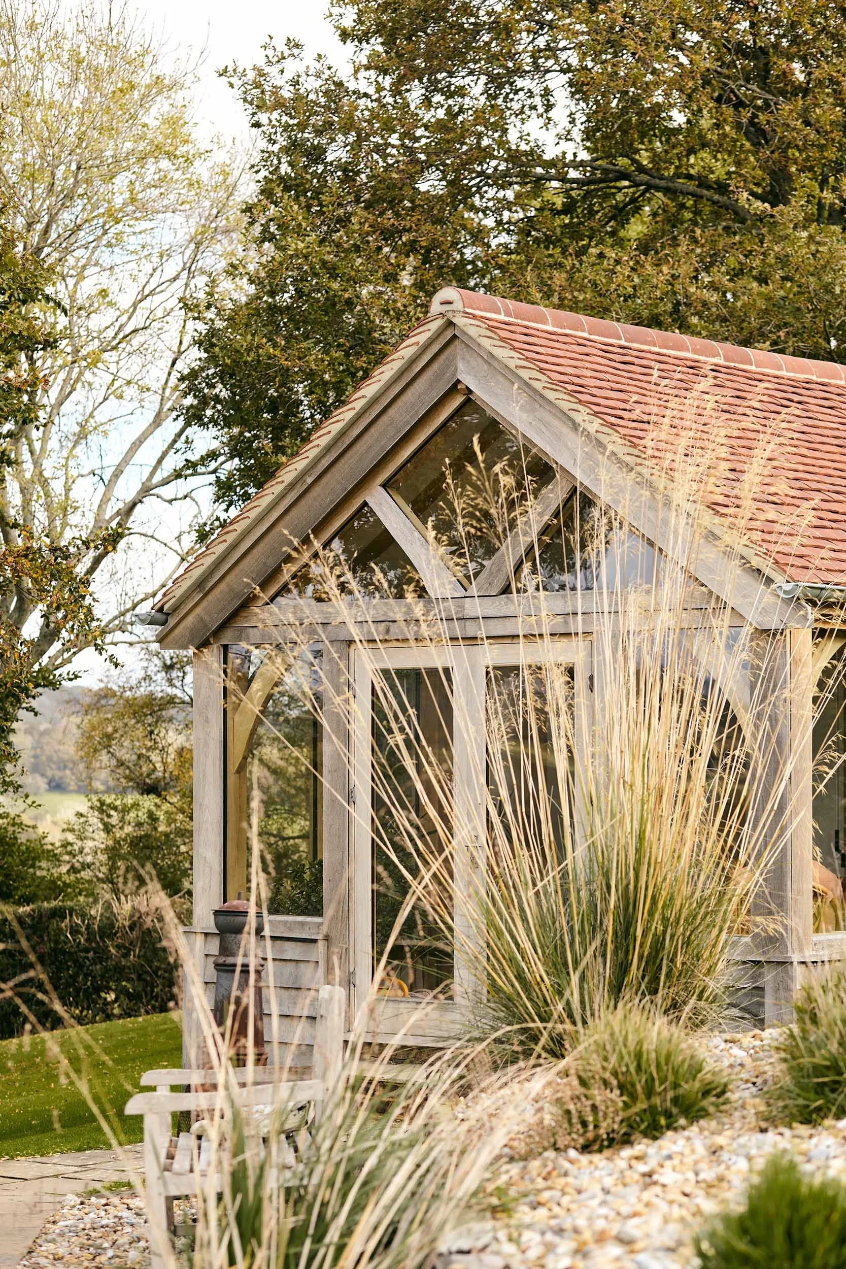 An oak framed garden room studio, with a red-tiled roof surrounded by tall grass and bushes in a garden setting.