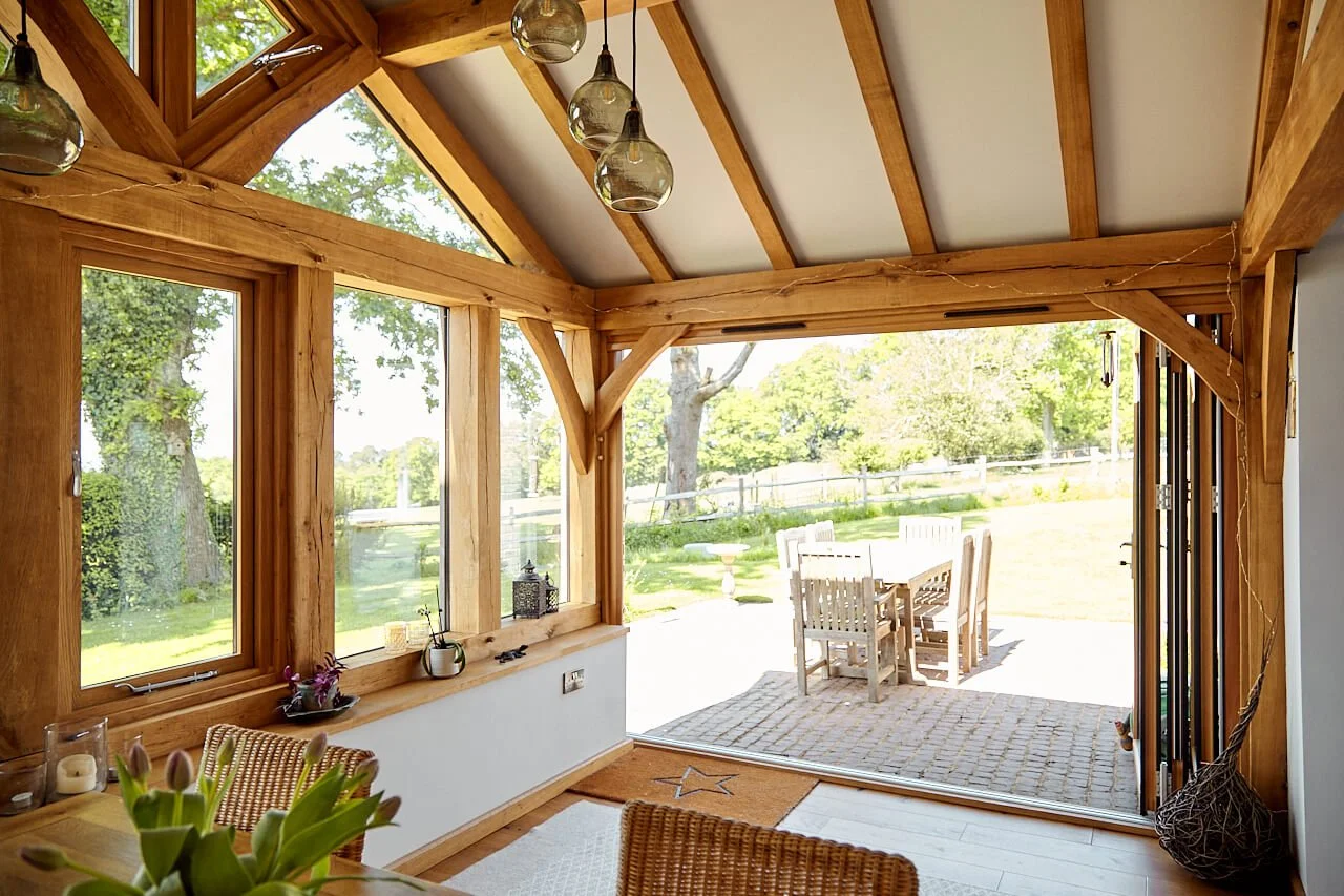 View from the interior of an oak framed sunroom extension, with large windows, and hanging glass light fixtures. An outdoor patio with a wooden table and chairs is visible through the open sliding door, with a lawn and trees in the background.