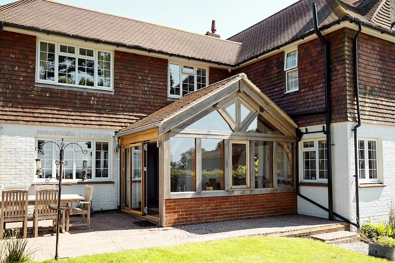 Back garden of a house featuring a brick and white painted exterior, large windows, and a newly constructed oak framed sunroom extension with a gable roof made of wood and glass panels.