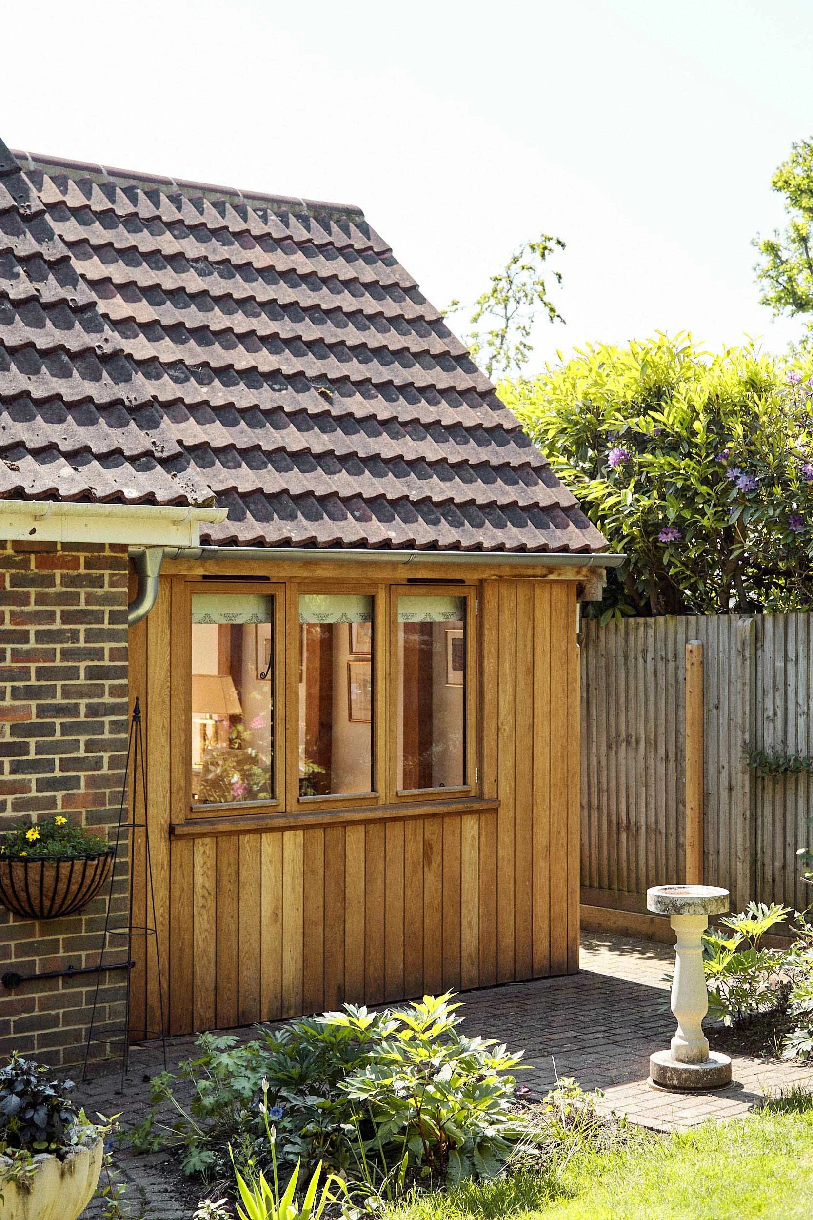 Side view of a brick house with an oak framed extension covered in oak channel cladding, a window, and a garden with plants and a birdbath.