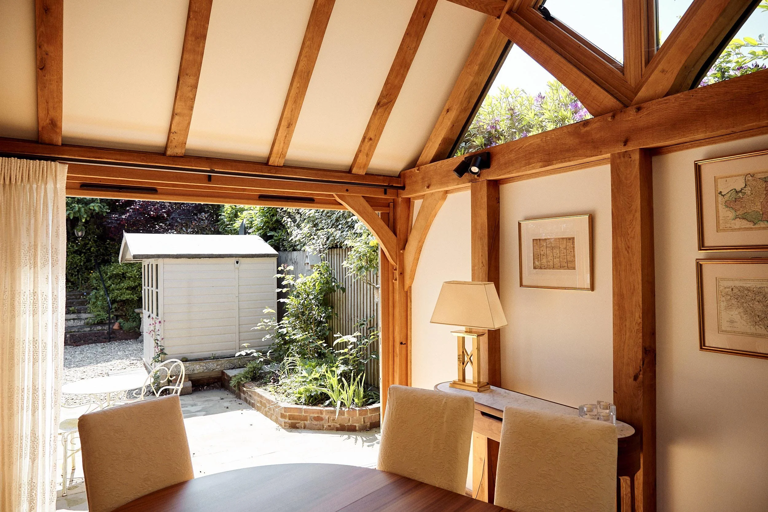 Sunlit dining area in an oak framed house extension, with wooden beams and a view of a garden shed and garden outside.