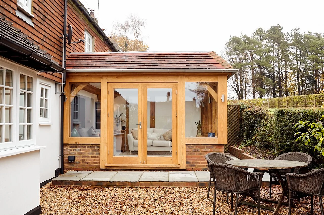 Back garden with a small patio and an oak framed sunroom extension to the house, outdoor table with chairs, bush and trees in the background.