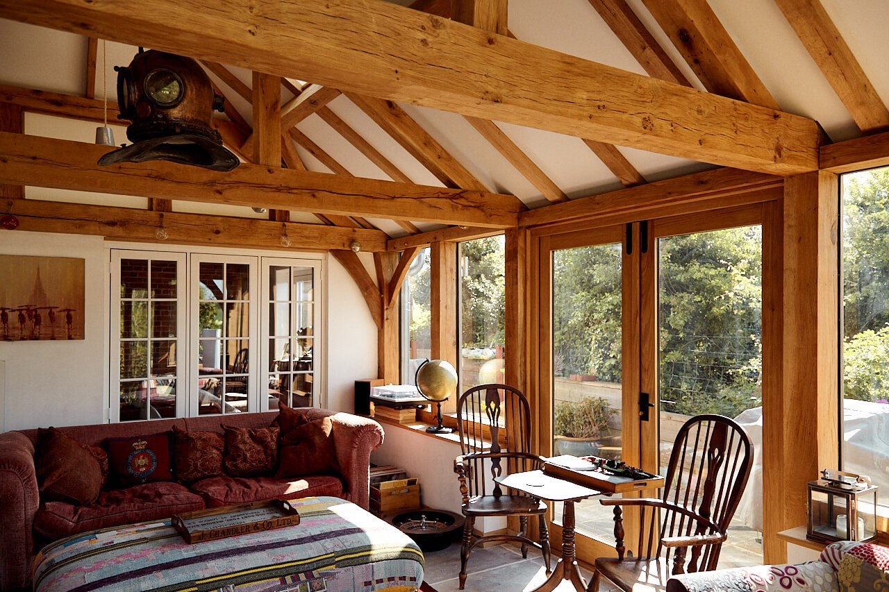 Sunlit living room in an oak framed house extension, with wooden beams, glass doors, a red sofa, a globe, wooden chairs, and various decorations. Large windows reveal a garden outside.