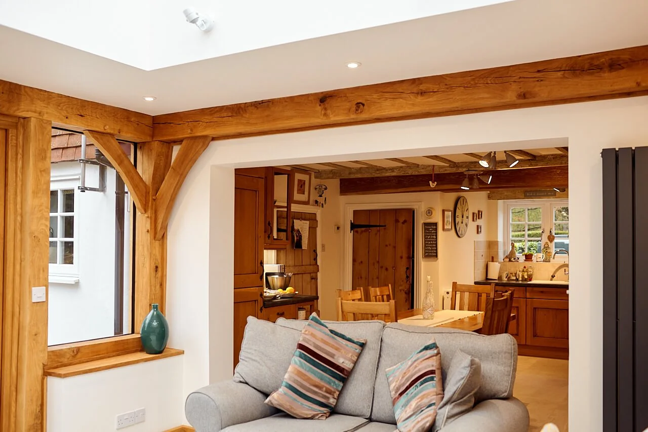 Living room in an oak framed house extension with wooden accents, a beige sofa with striped pillows, and a view into a kitchen and dining area with wooden cabinets and furniture.