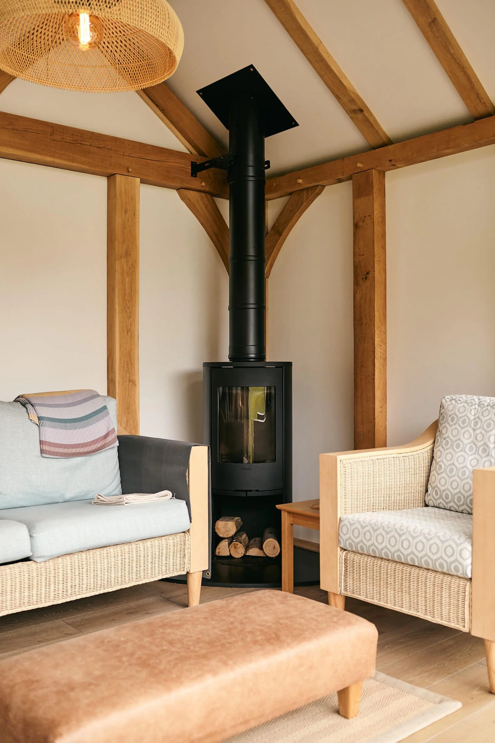 Living space in an oak framed garden studio, with a wood stove, a light coloured wicker sofa with pale cushions, an armchair with patterned cushions, a wooden side table, and a brown upholstered ottoman. Visible wooden beams and a rattan ceiling ligh