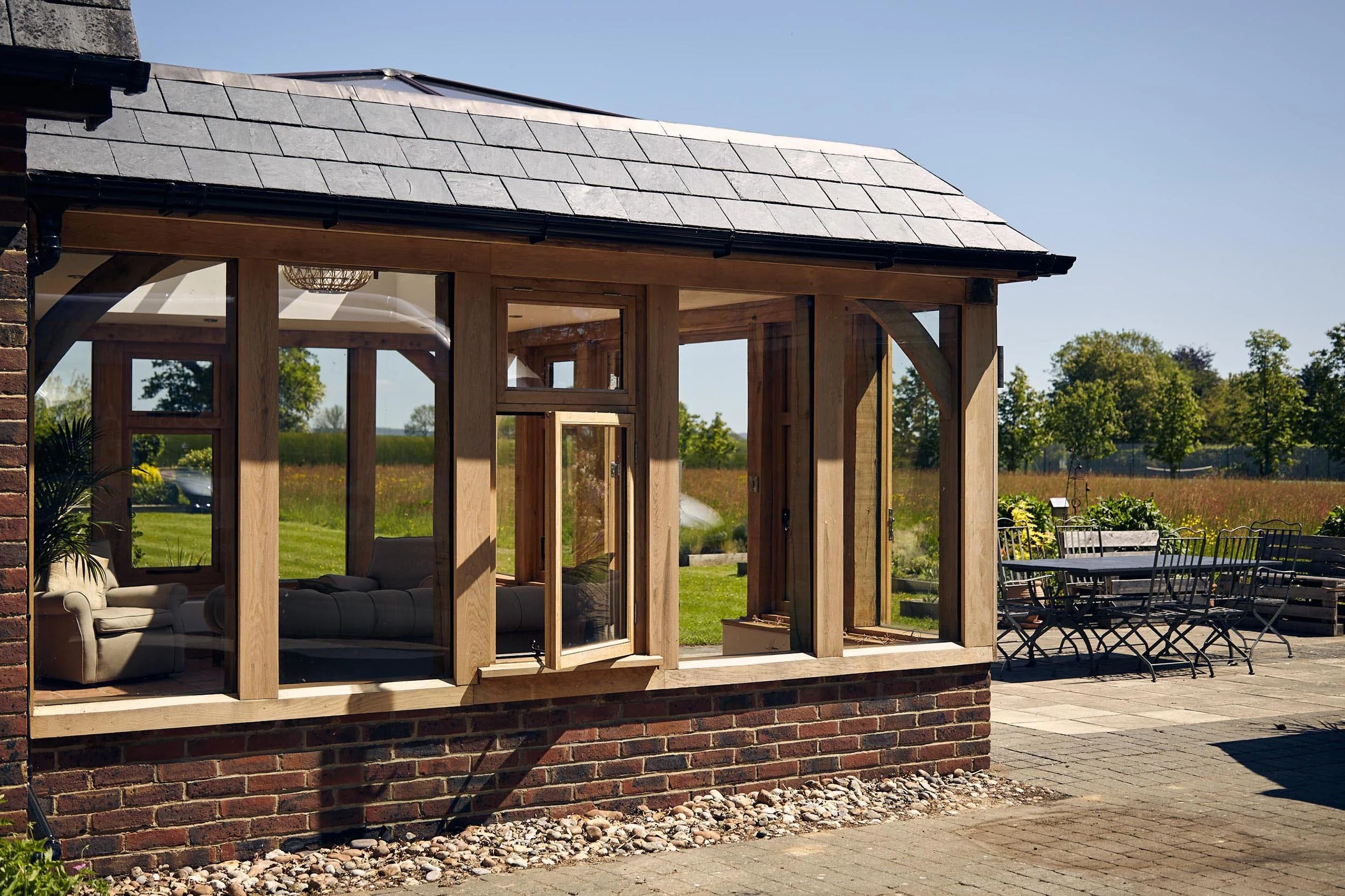 A brick house with an oak framed orangery extension, surrounded by an outdoor patio with metal chairs and tables, in a rural setting with green trees and open field in the background.