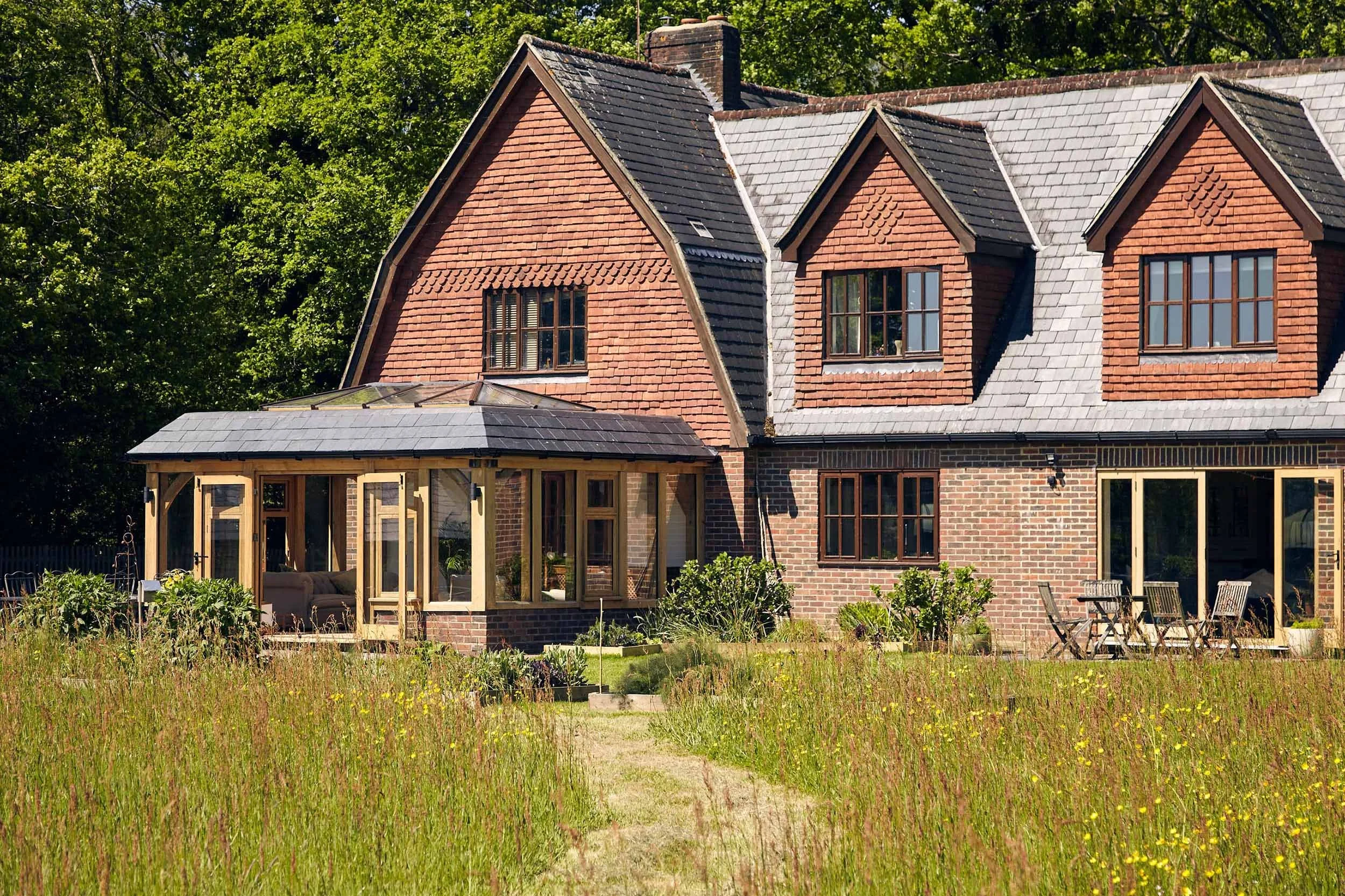 A two-story brick house with dormer windows and an oak framed orangery extension, set in a grassy field with trees in the background.