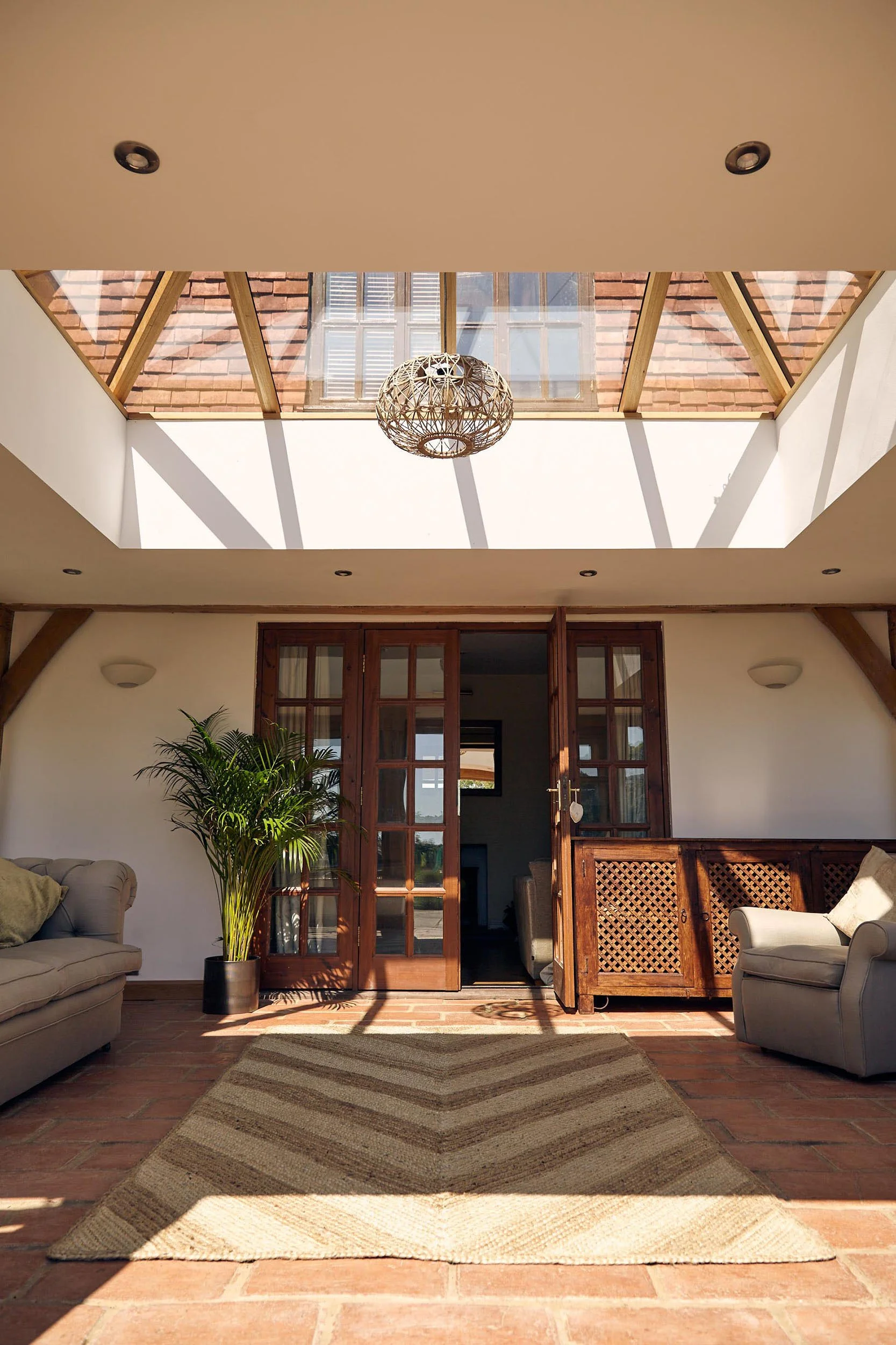 Living room in an oak framed orangery with a skylight, wooden furniture, a beige carpet, and two sofas, one beige and one gray, with a large potted plant next to the door.